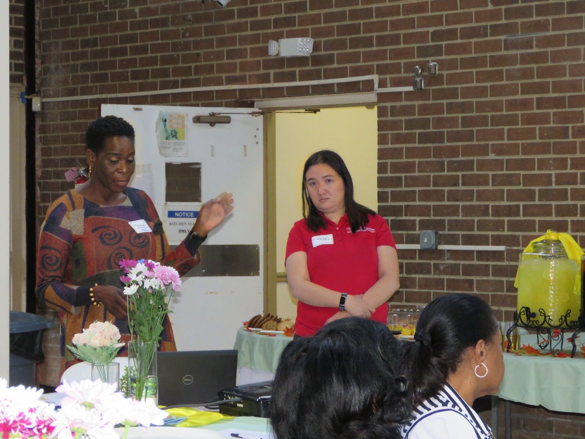 Two women at a table; one speaks, gesturing. A table with snacks and flowers. Brick wall background.