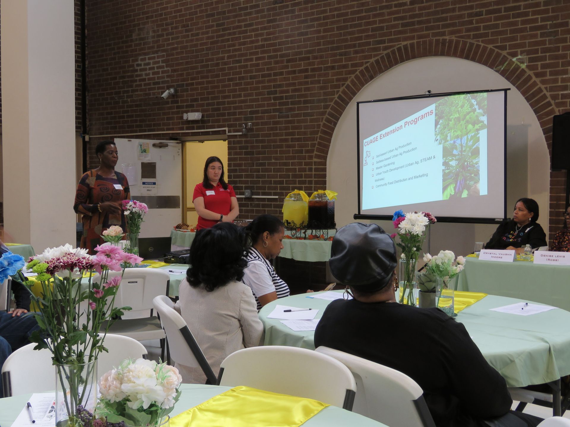 People at a presentation; speakers by screen and audience at tables in a hall with brick walls.