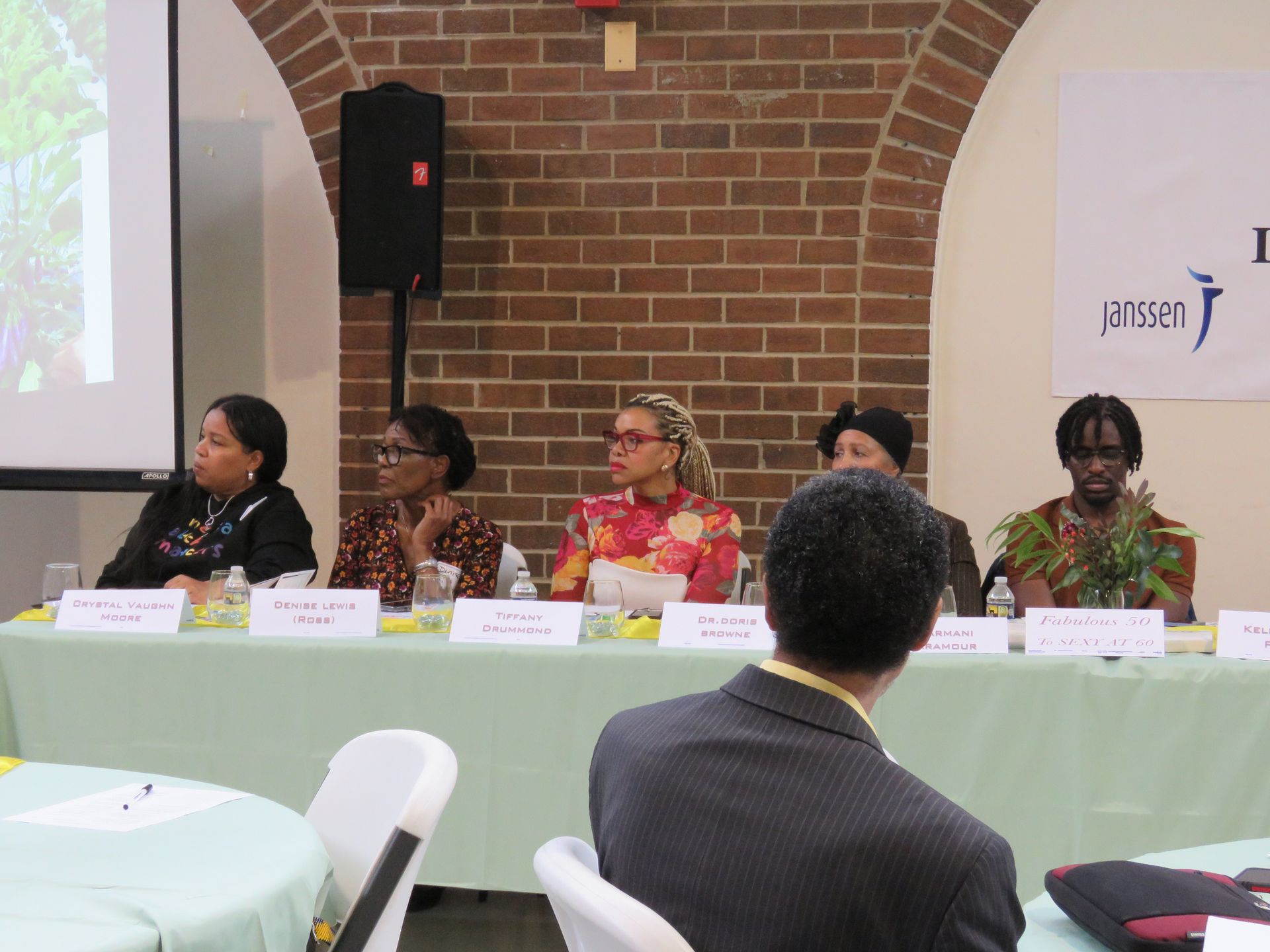 Panel of five people at a conference table, facing forward. Audience member in foreground. Brick wall background.