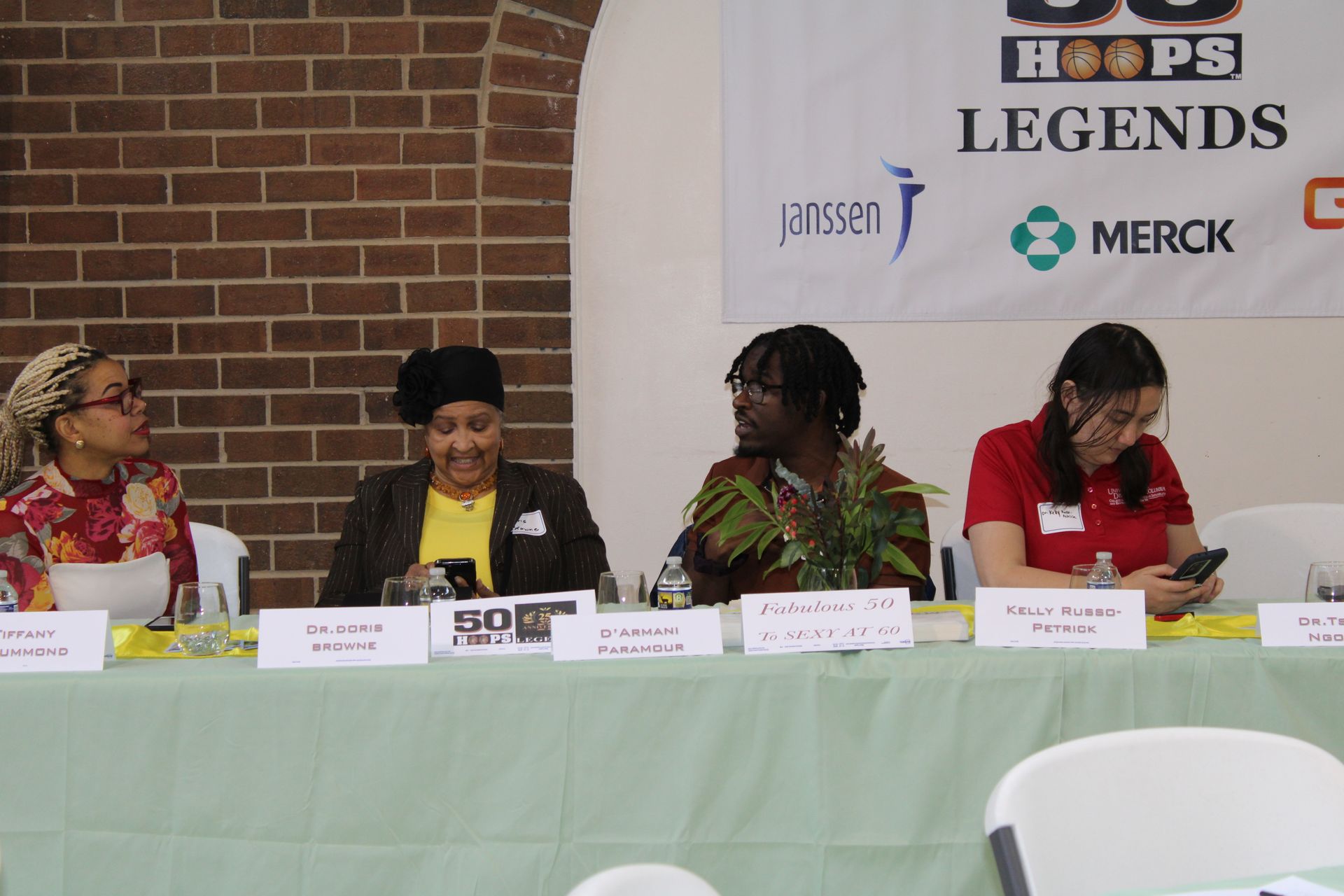 Panel of four people seated behind a table with signs. Event banner, brick wall background.