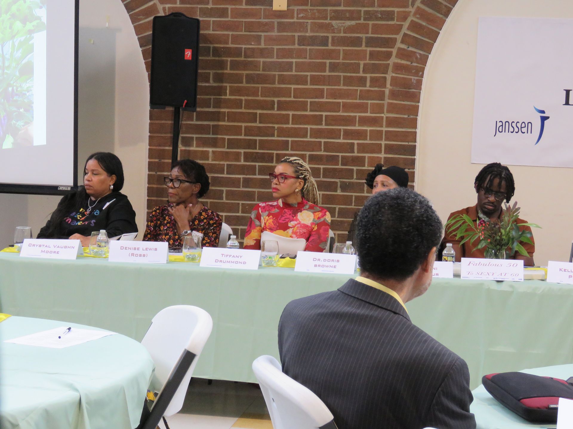Panel of six people seated at a table, speaking at an event in front of a brick wall, logo visible.
