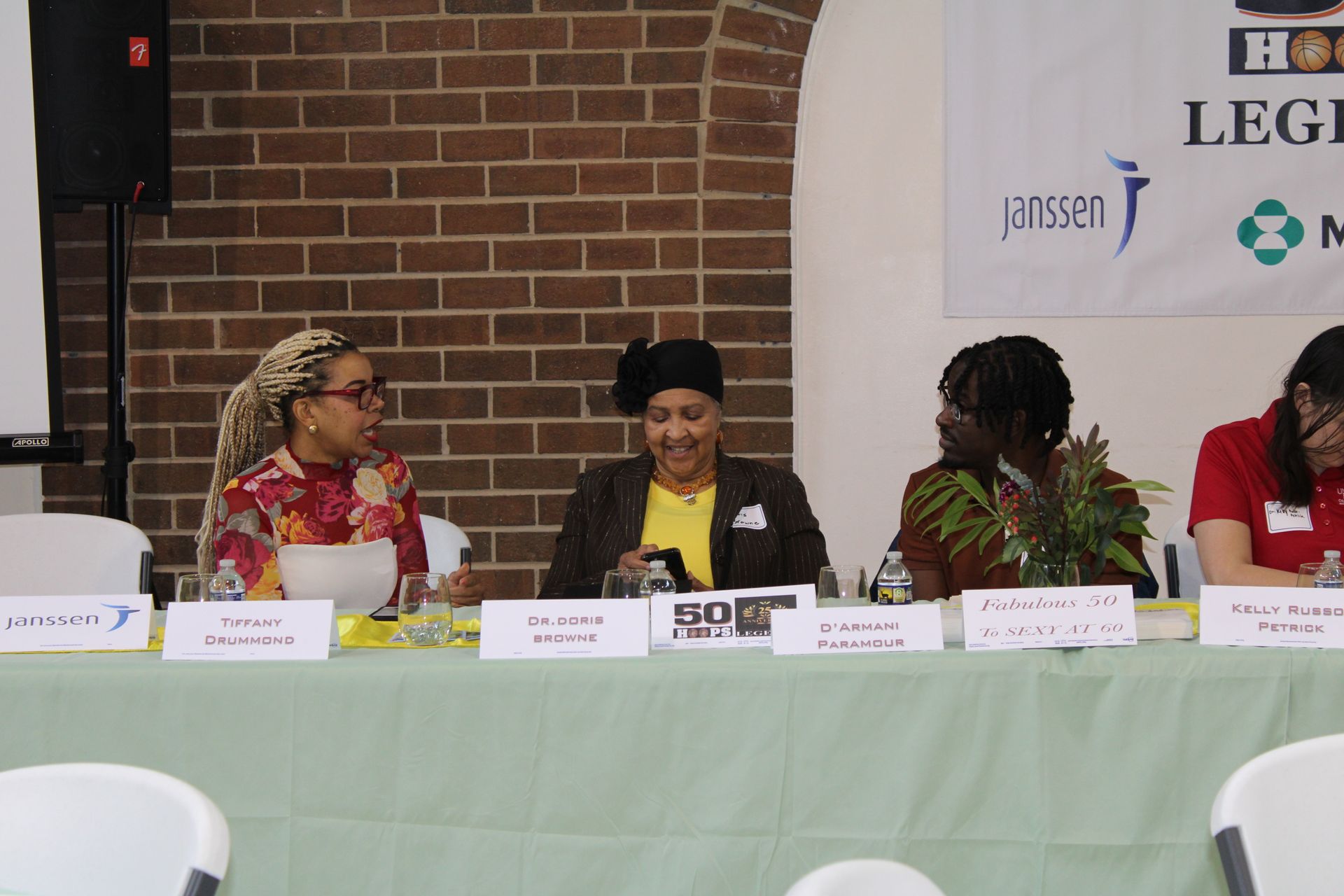 Panel of four people seated at a table with name placards in front of them, banner in the background.