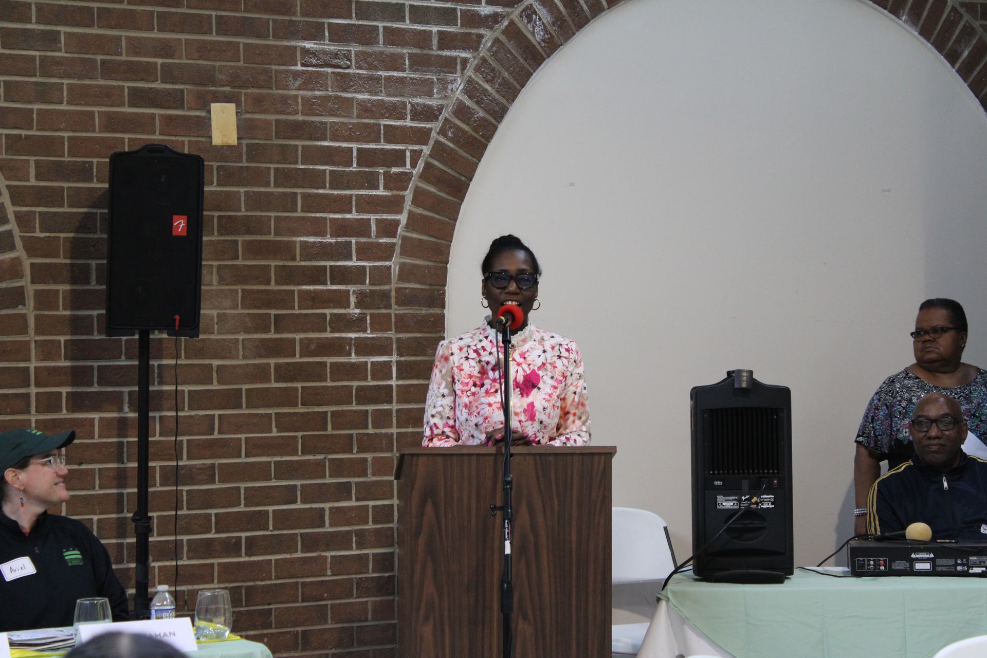 Woman speaking at a podium with microphone, people seated nearby, brick wall backdrop.