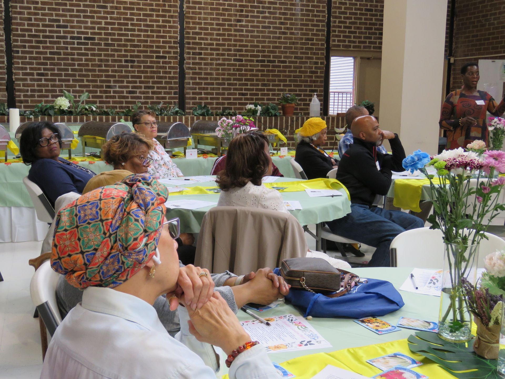 Attendees listen to a presentation at a flower-decorated event in a brick-walled room.