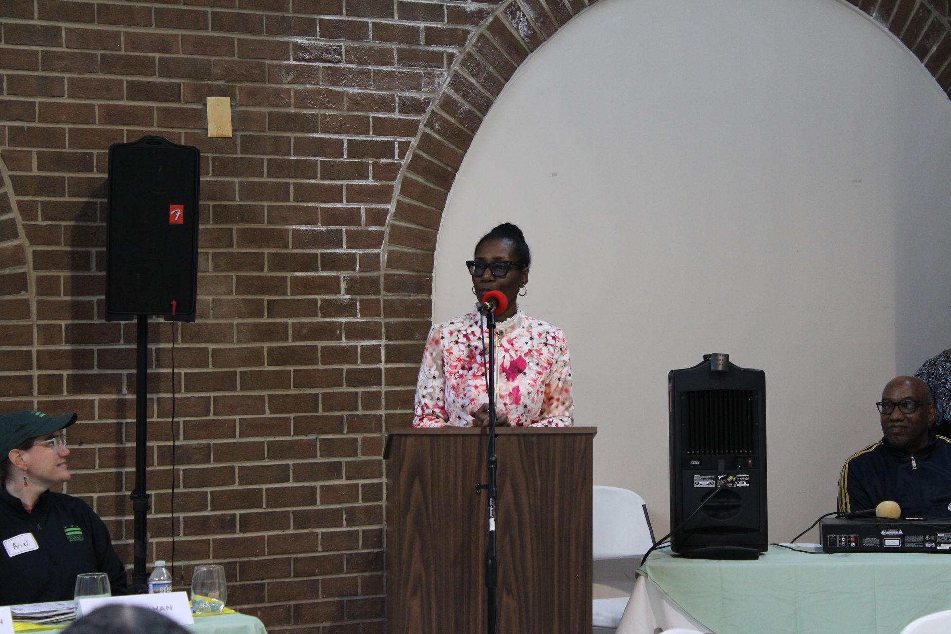 Woman speaking at a podium, facing a crowd. Brick wall backdrop. Speaker next to a speaker and a police officer.