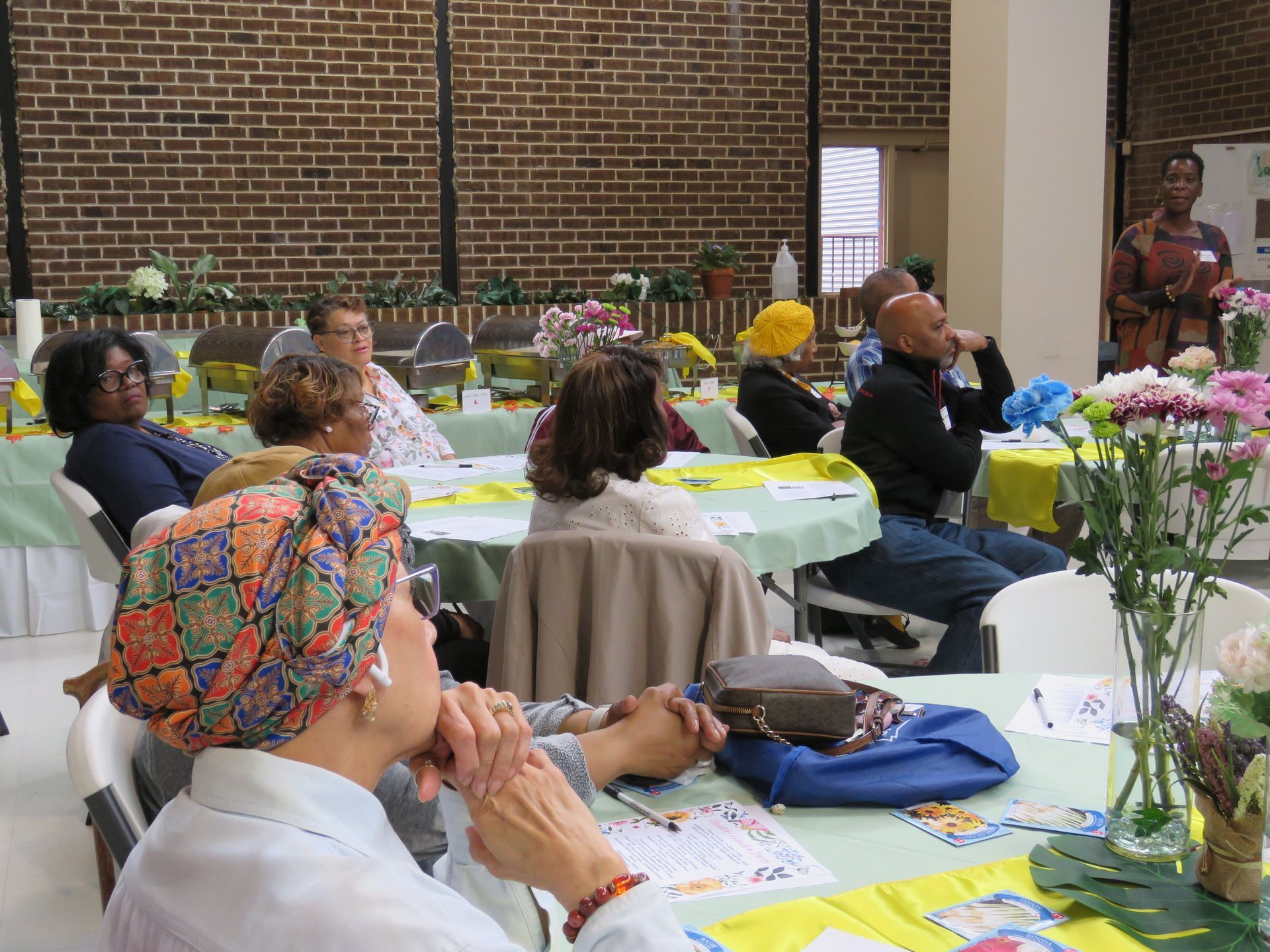 People seated at tables, listening to a speaker in a room with a brick wall and flowers.