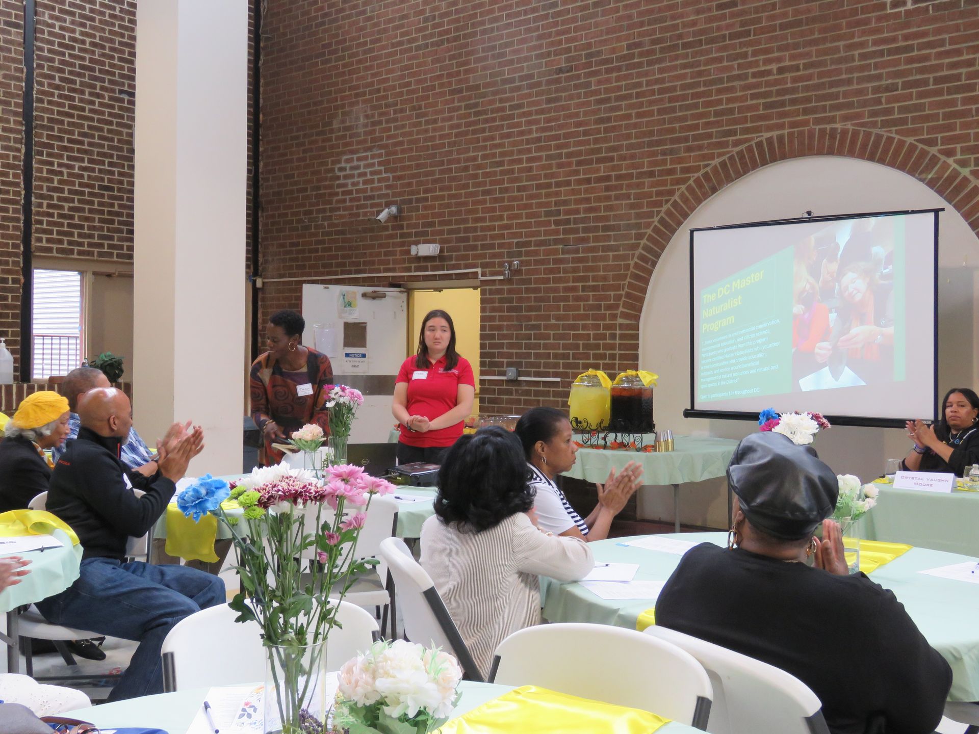A woman presenting to a seated audience at an event in a brick-walled hall.