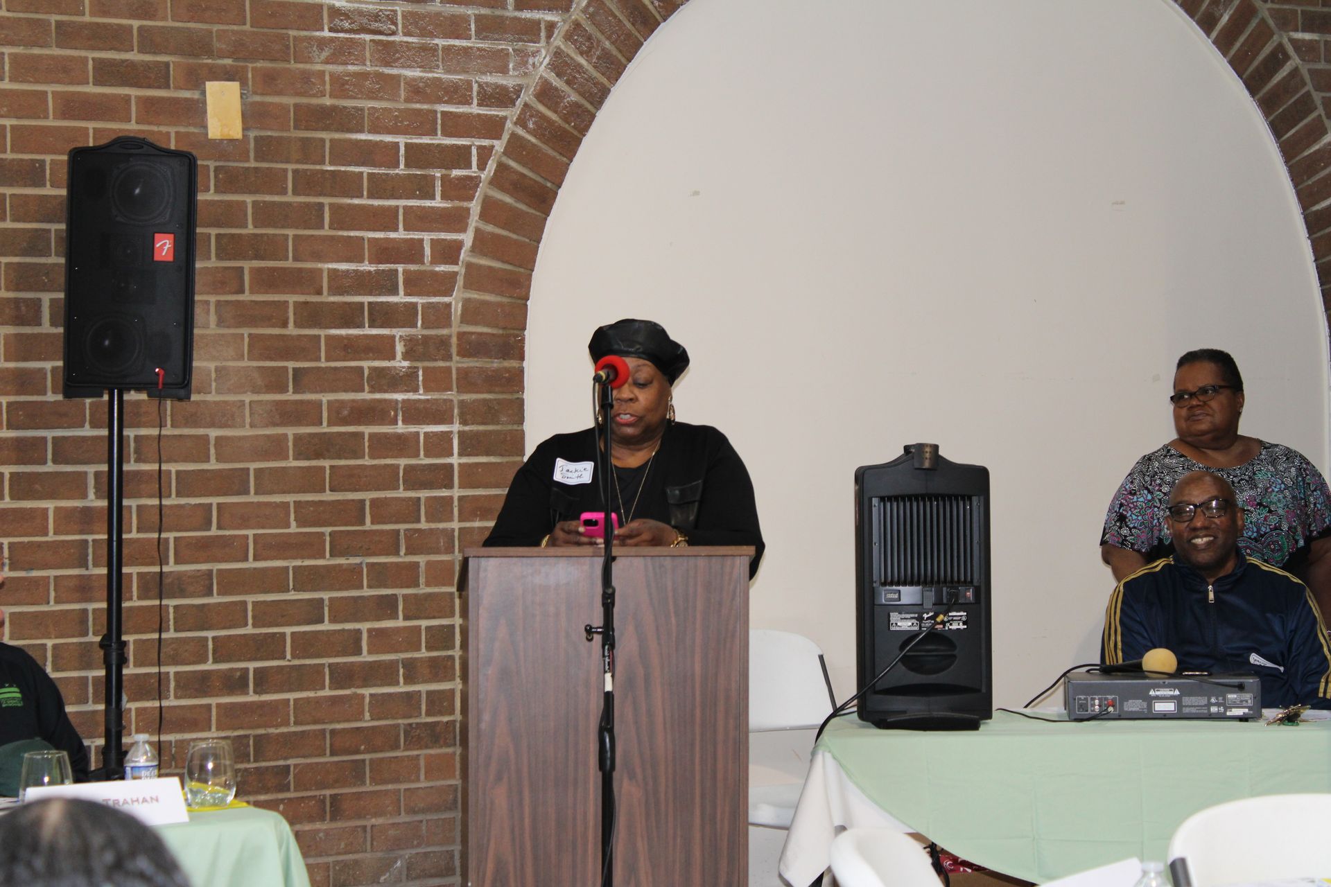 Woman speaking at a podium, two others seated nearby, in a brick-walled room.