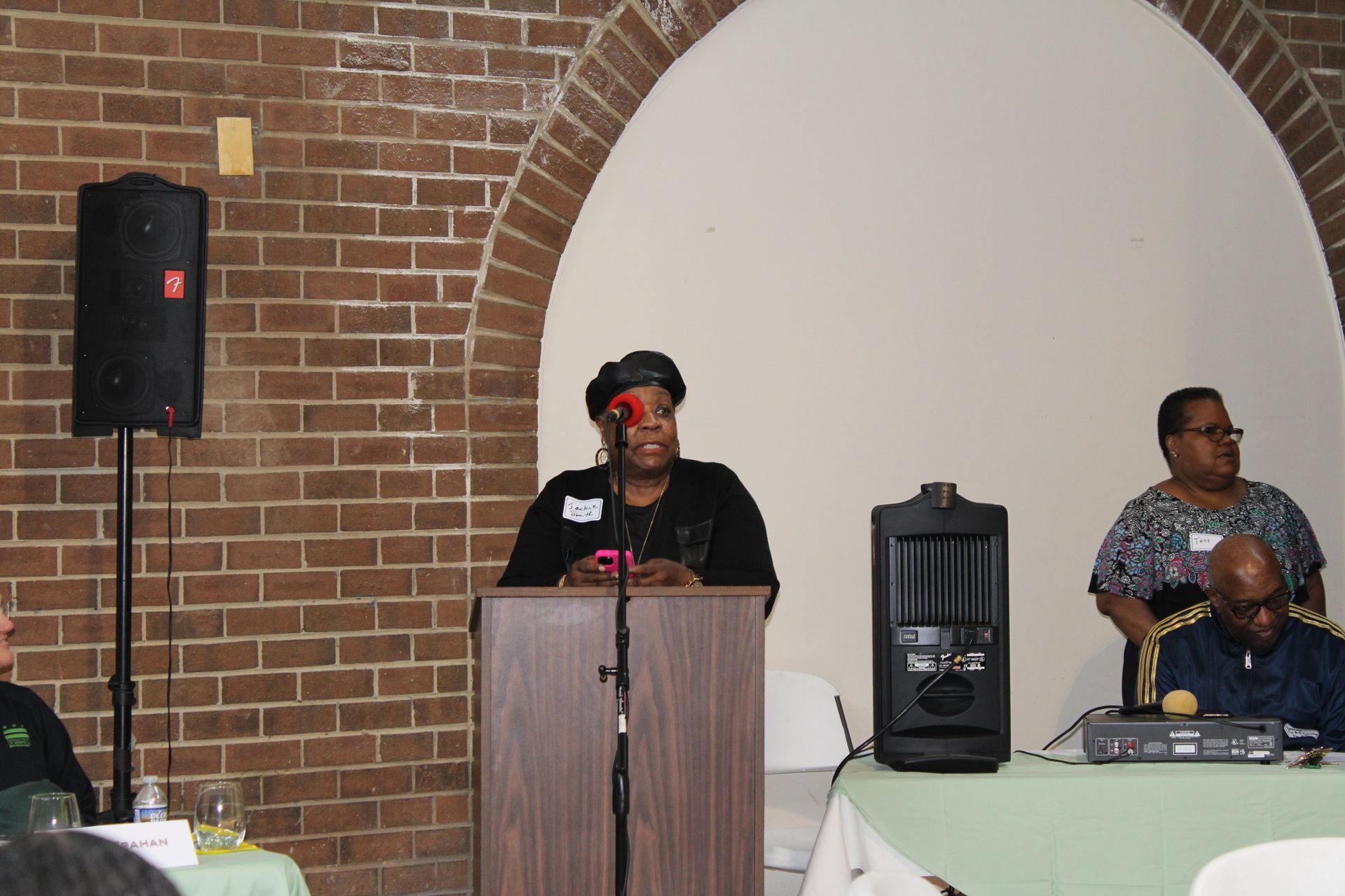 Woman speaking at a podium in front of a brick wall and a seated audience, with audio equipment.