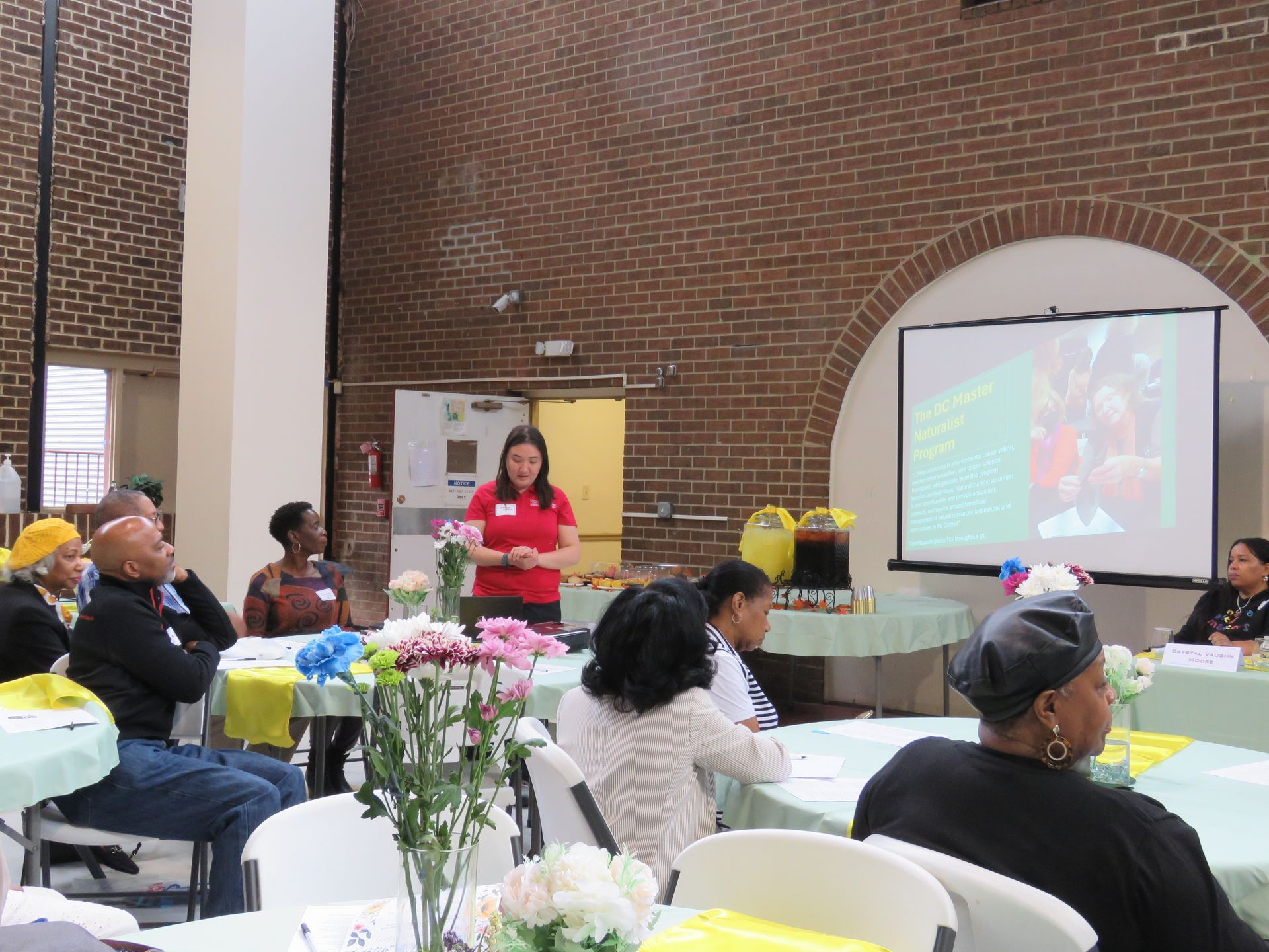 A presenter addresses seated audience at a meeting. Projector screen shows a slide. Tables are set with flowers.