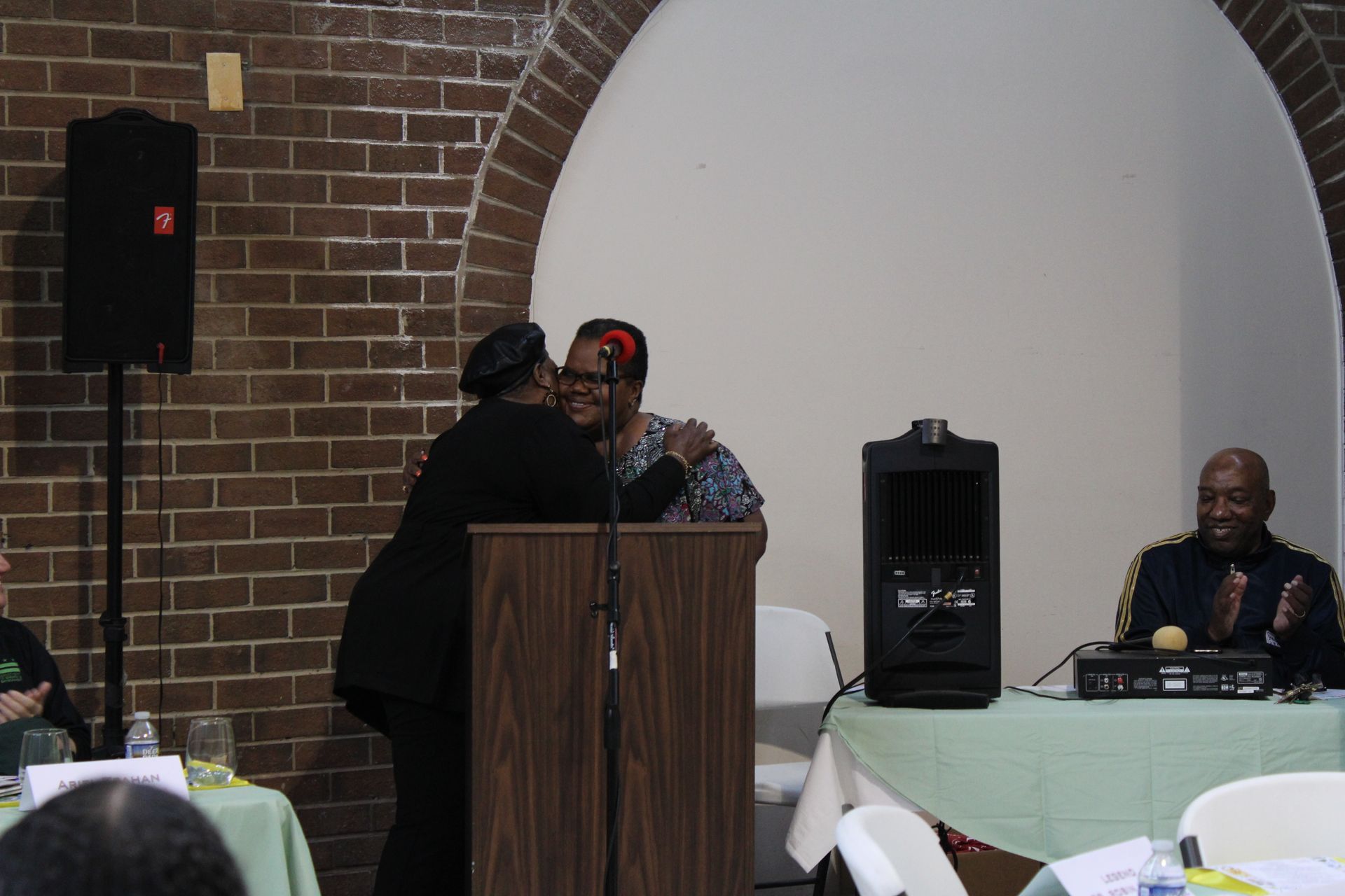 Two women embrace at a podium during an event. A man smiles at a table. Brick wall in background.