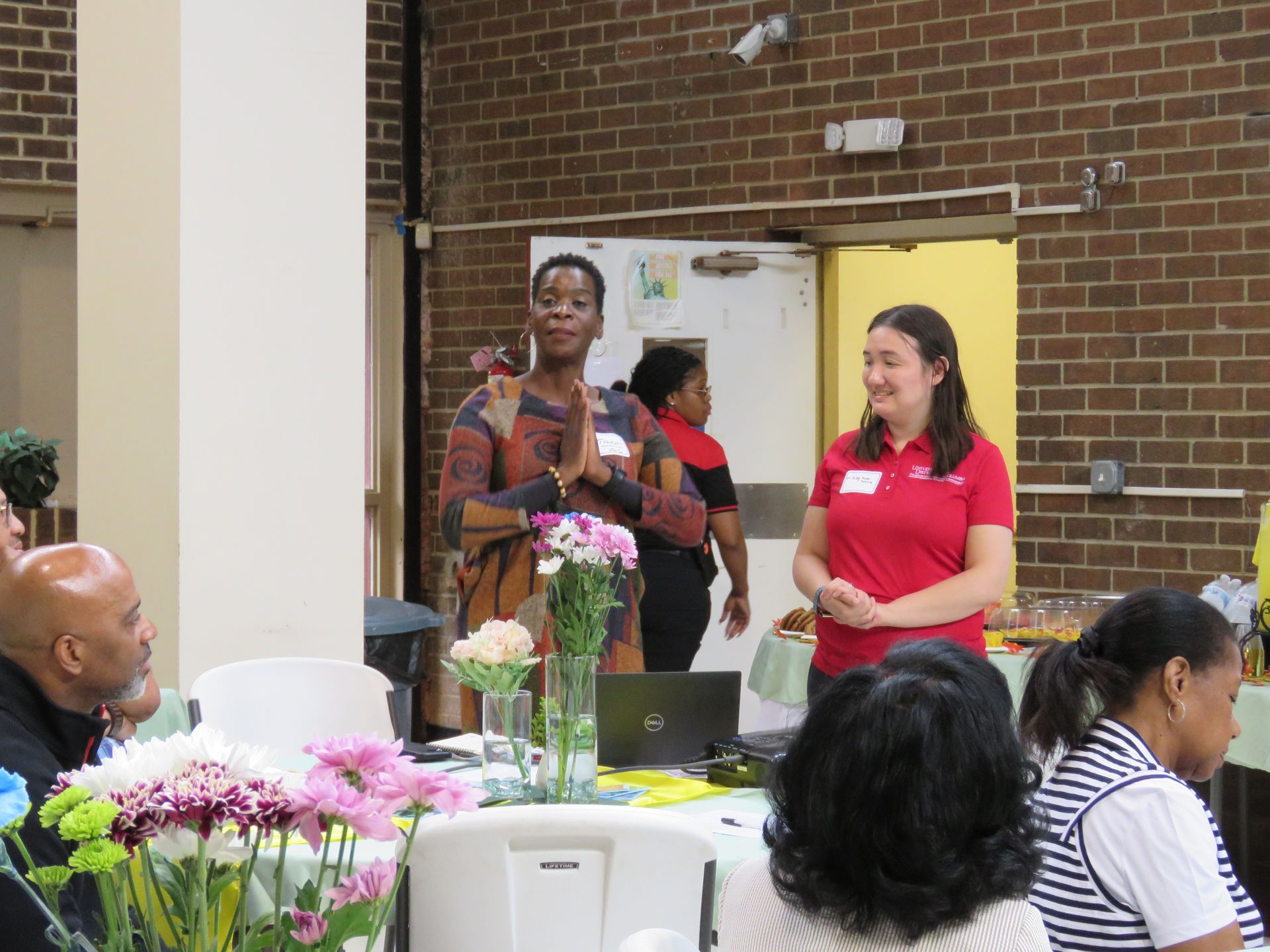 Woman speaking at an event, another woman stands nearby. Tables with flowers and people in a brick-walled room.