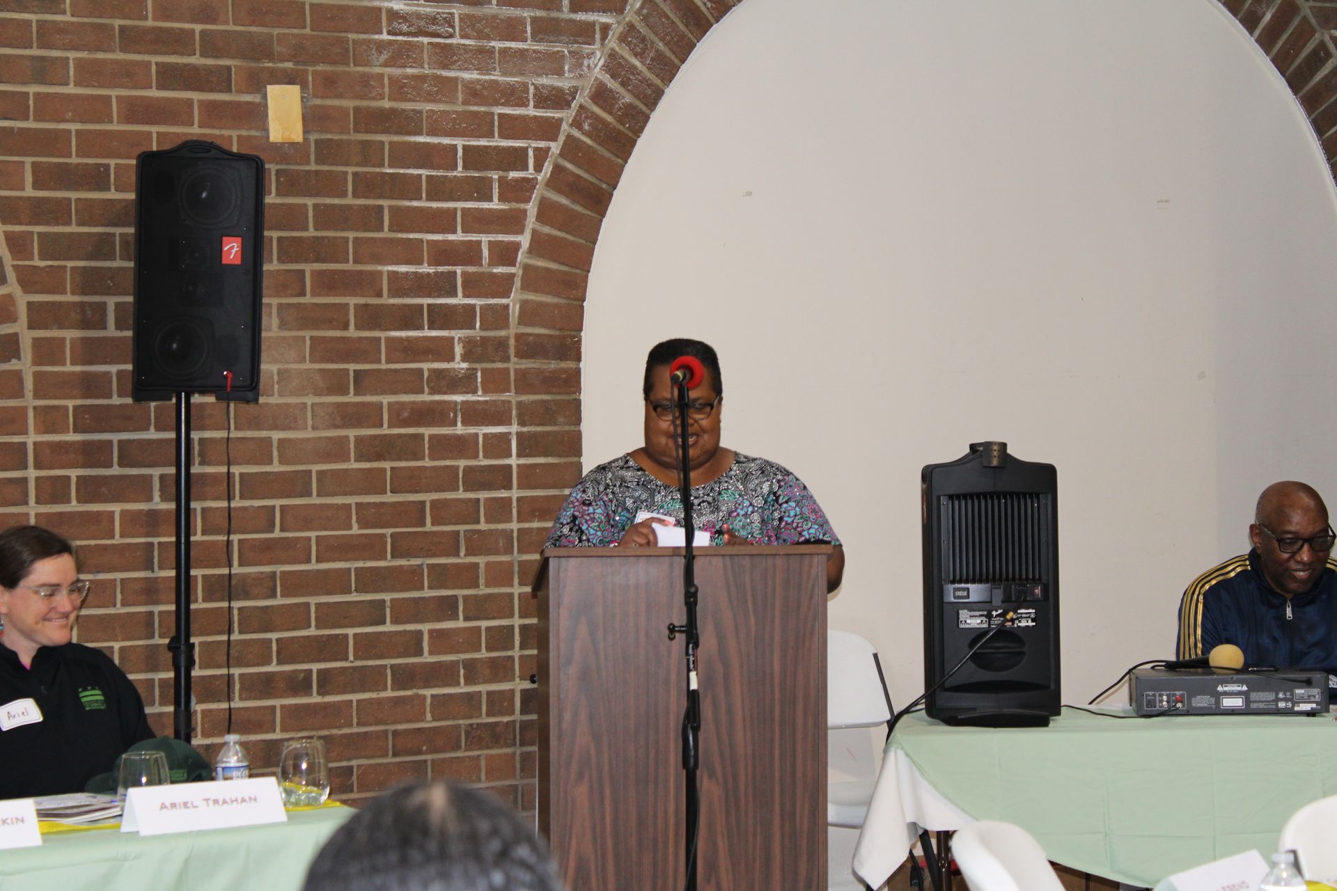 Woman speaking at podium during event; others seated at table with brick wall backdrop.