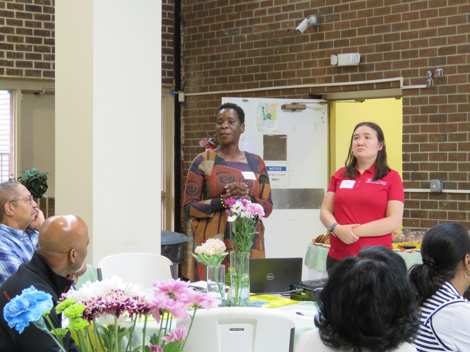 Two women presenting at a table with flowers. One speaks, the other stands beside. Audience in the background.