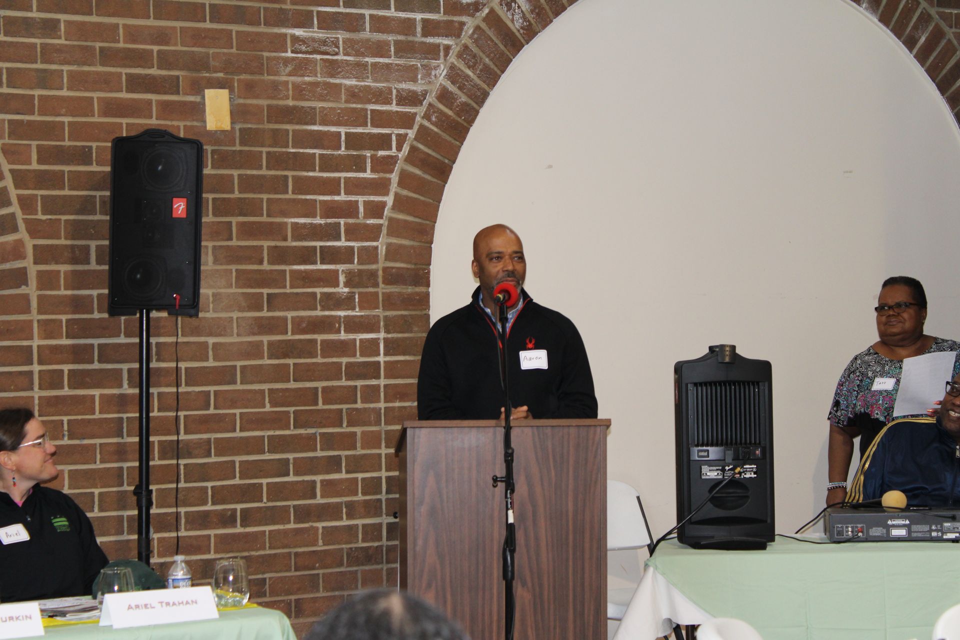 Man speaking at a podium with two others at a table in front of brick wall. Speaker with microphone and speaker.