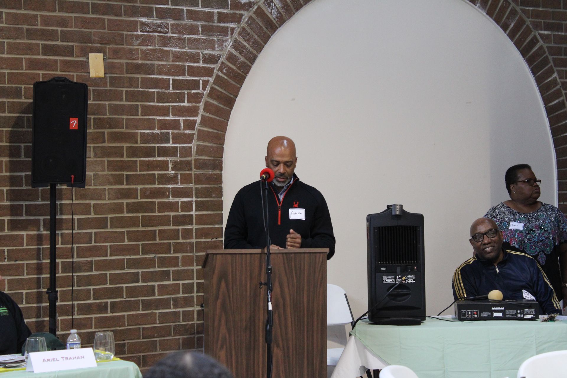 Man speaking at a podium in a brick-walled room; two people watch.