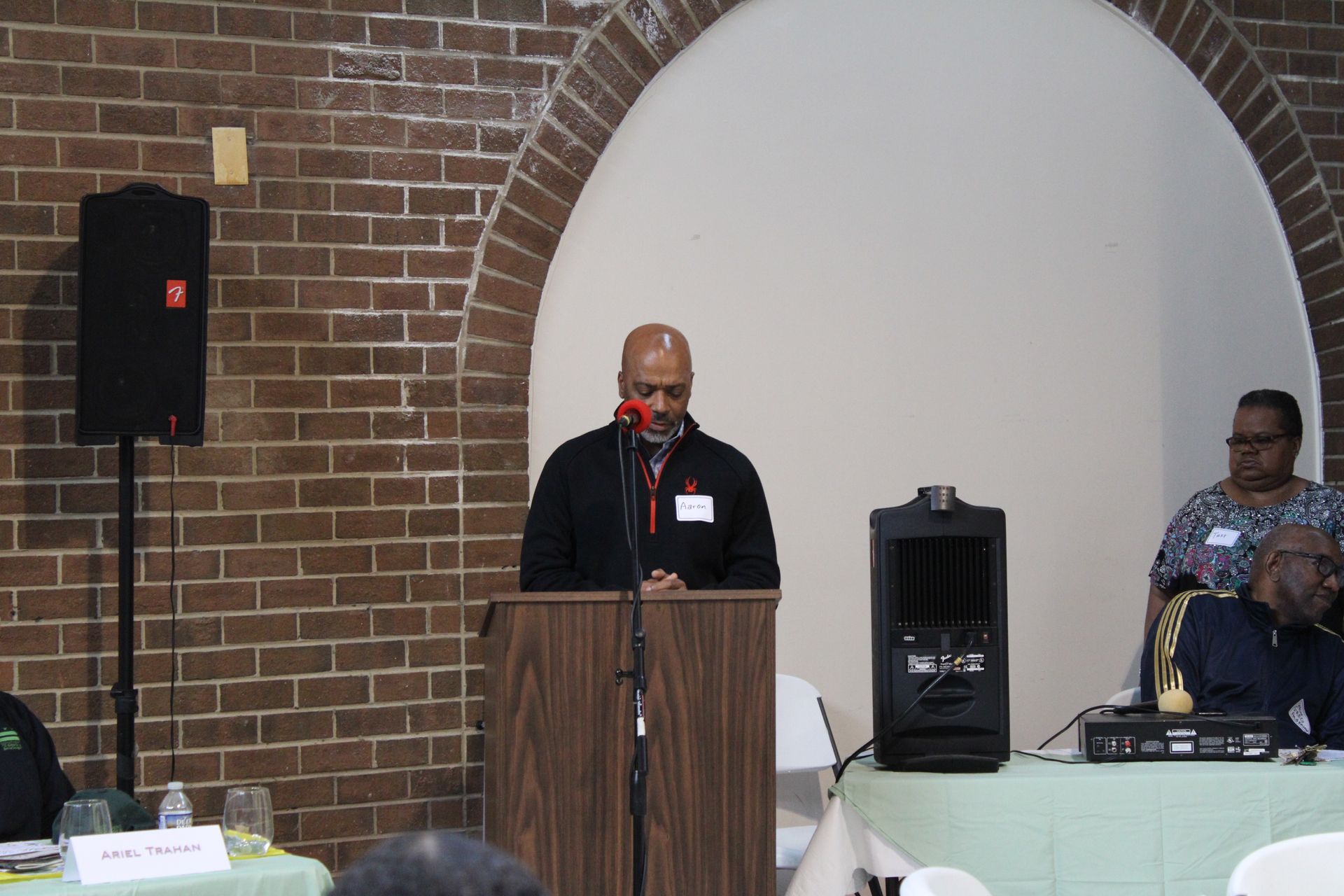 Man speaking at a podium in a brick-walled room with a speaker. Others sit nearby.