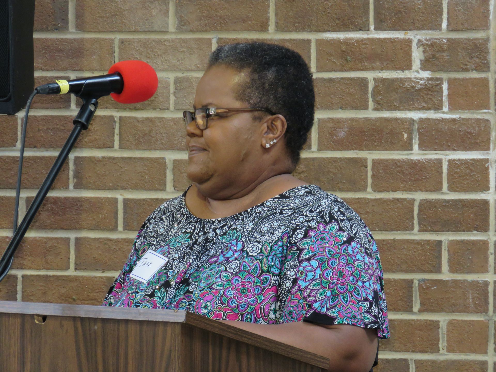 Woman speaking at a podium, wearing glasses and a floral print top, with a microphone in front of her. Brick wall background.