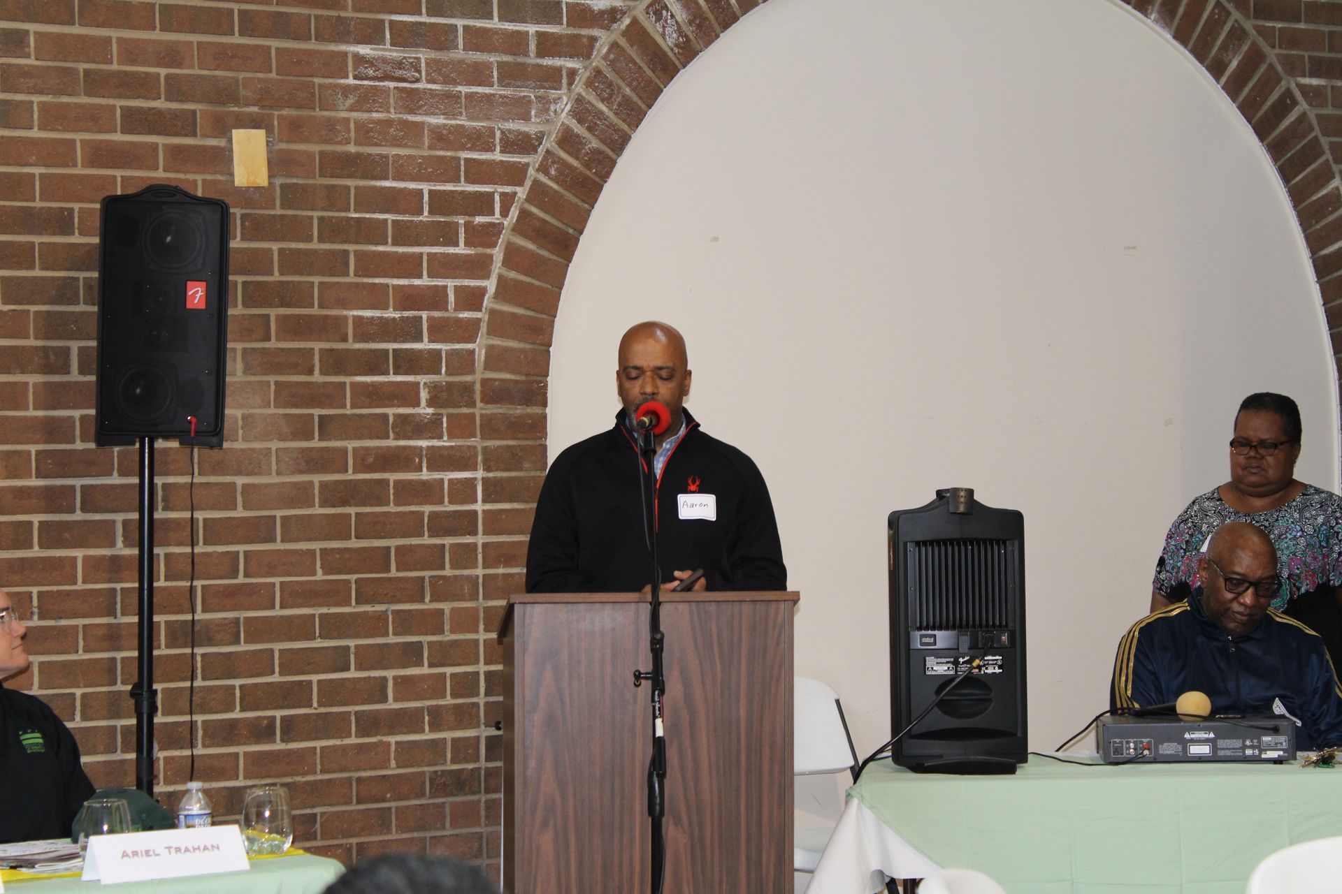 Man speaking at a podium under an arched opening in front of a brick wall, with audience members visible.