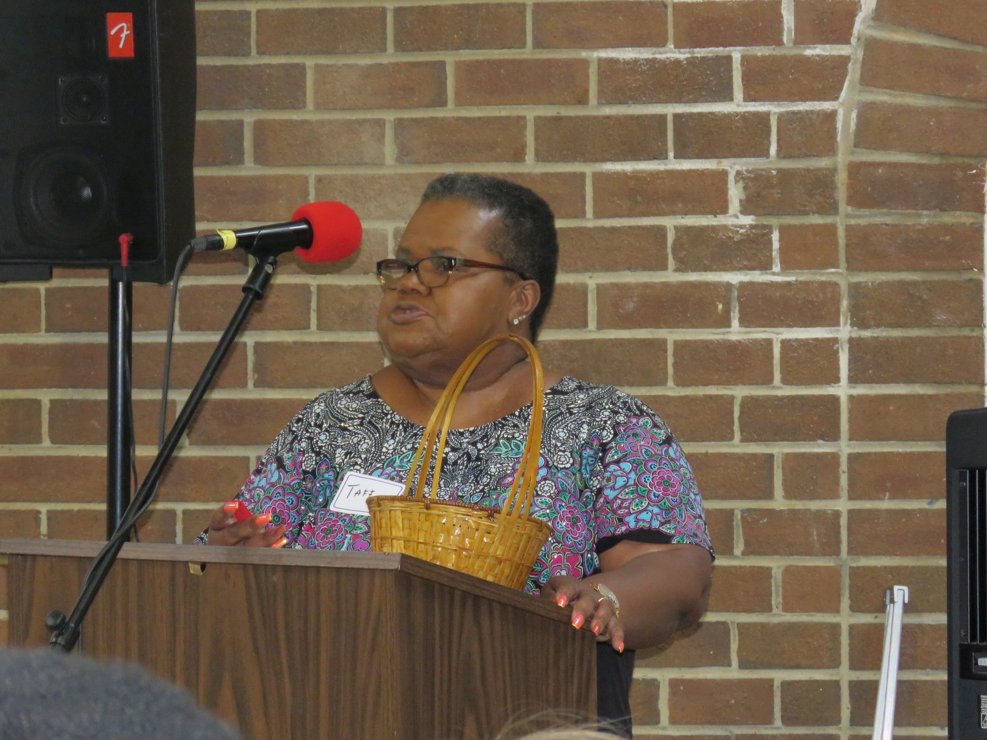 Woman speaking at a podium, holding a basket, microphone, brick wall background.