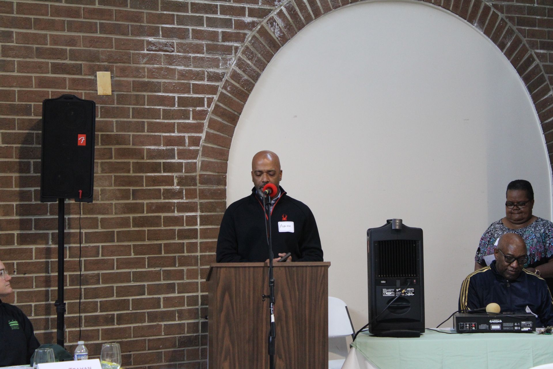 Man speaking at a podium with microphone. Brick wall, arched alcove, and two other people present.