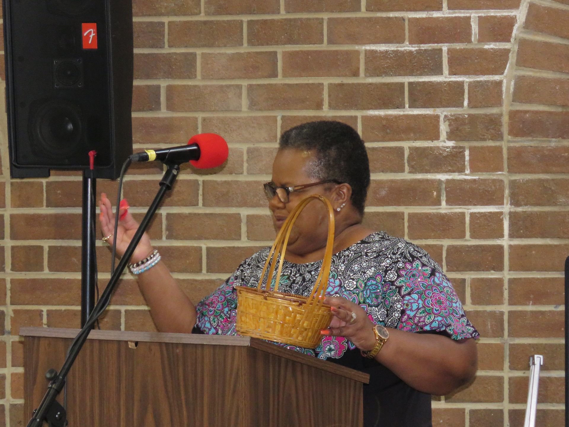 Woman at podium with microphone, holding a wicker basket, speaking near a brick wall.