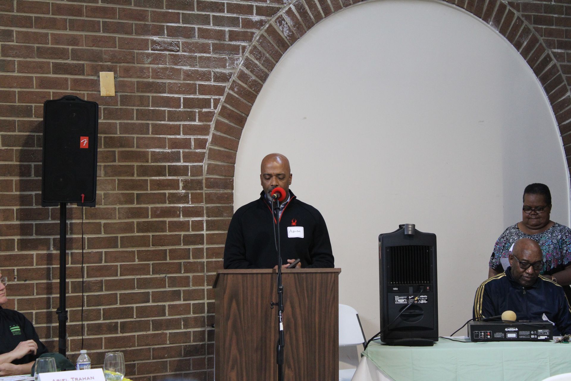 Man speaking at a podium, brick wall background, another person at a table, arched doorway.