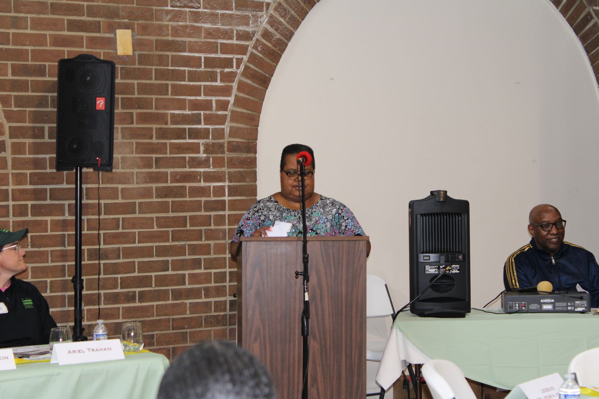 Woman speaking at podium, two others seated. Panel discussion in room with brick wall.