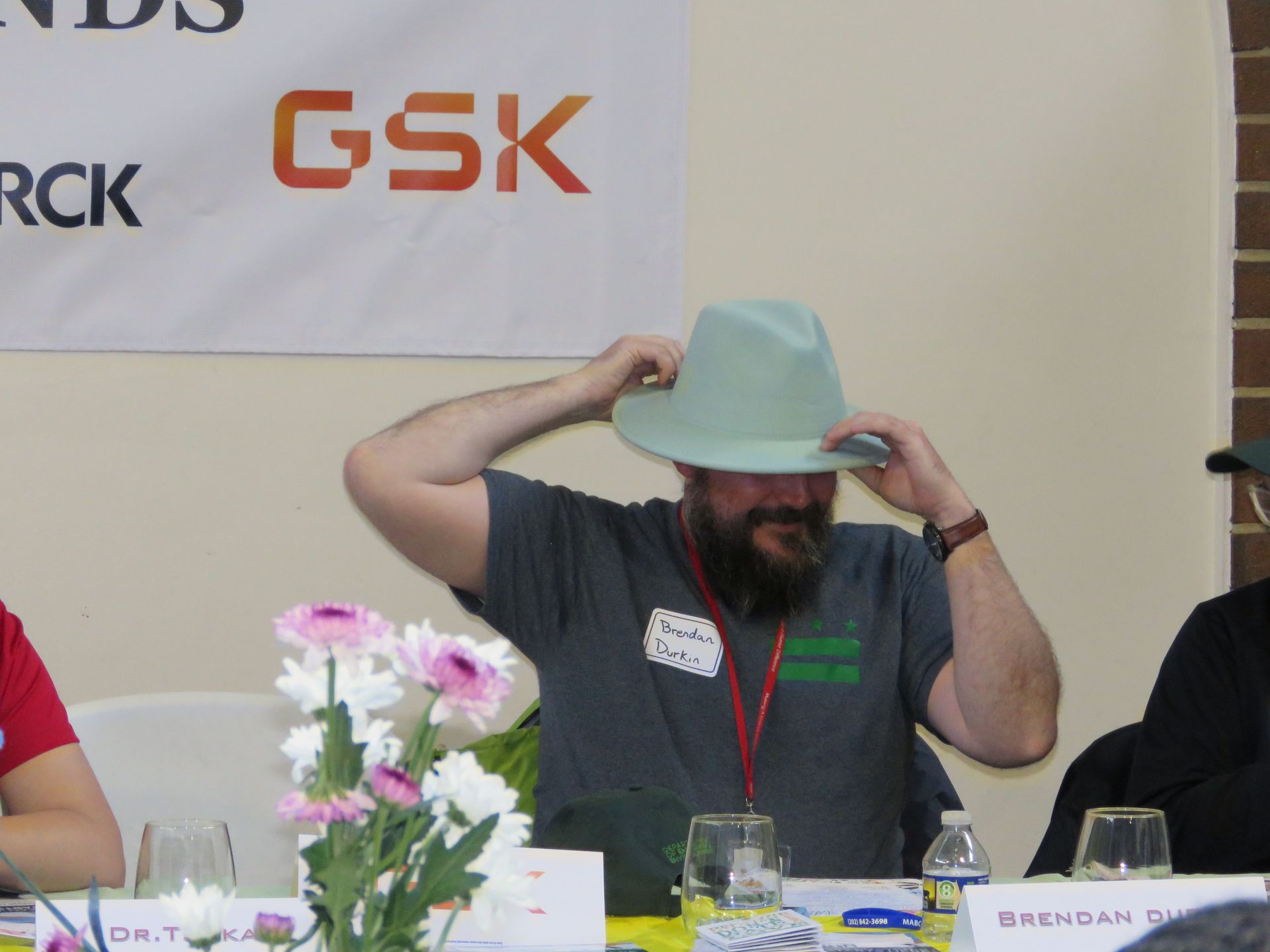 Man with beard adjusting a light green hat at a table with flowers. GSK logo in the background.