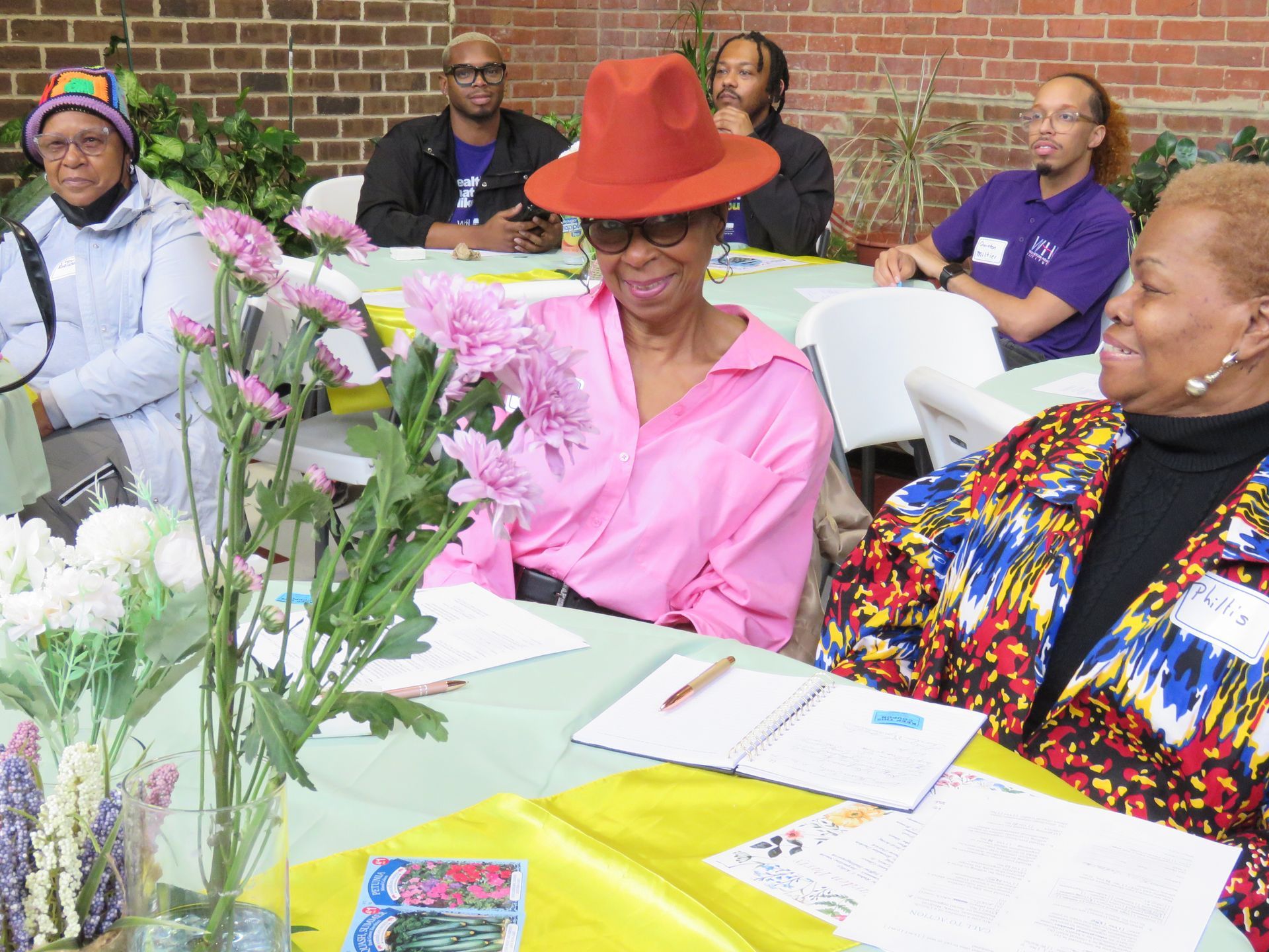 Group of people seated at tables, one woman in a pink shirt and hat examines flowers.