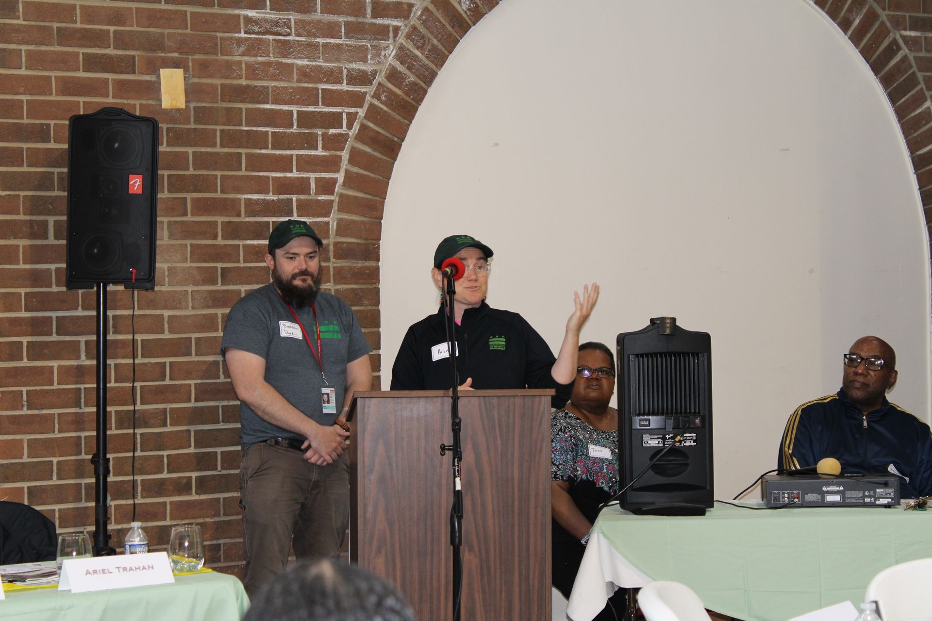 Two people speak at a podium, one gesturing. Others sit at tables in a room with a brick wall and archway.