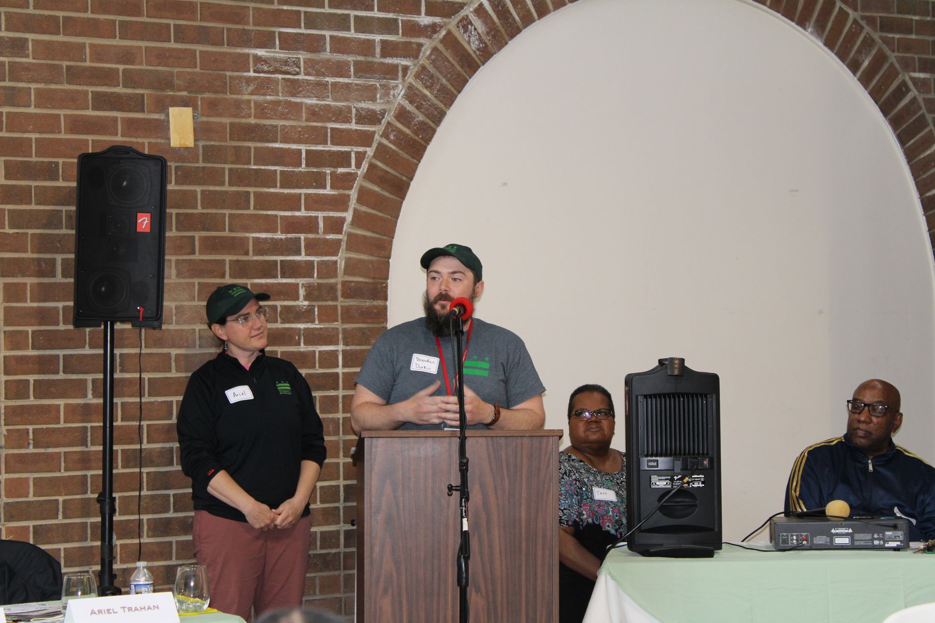 A man speaks from a podium with a woman beside him, speakers and brick background.