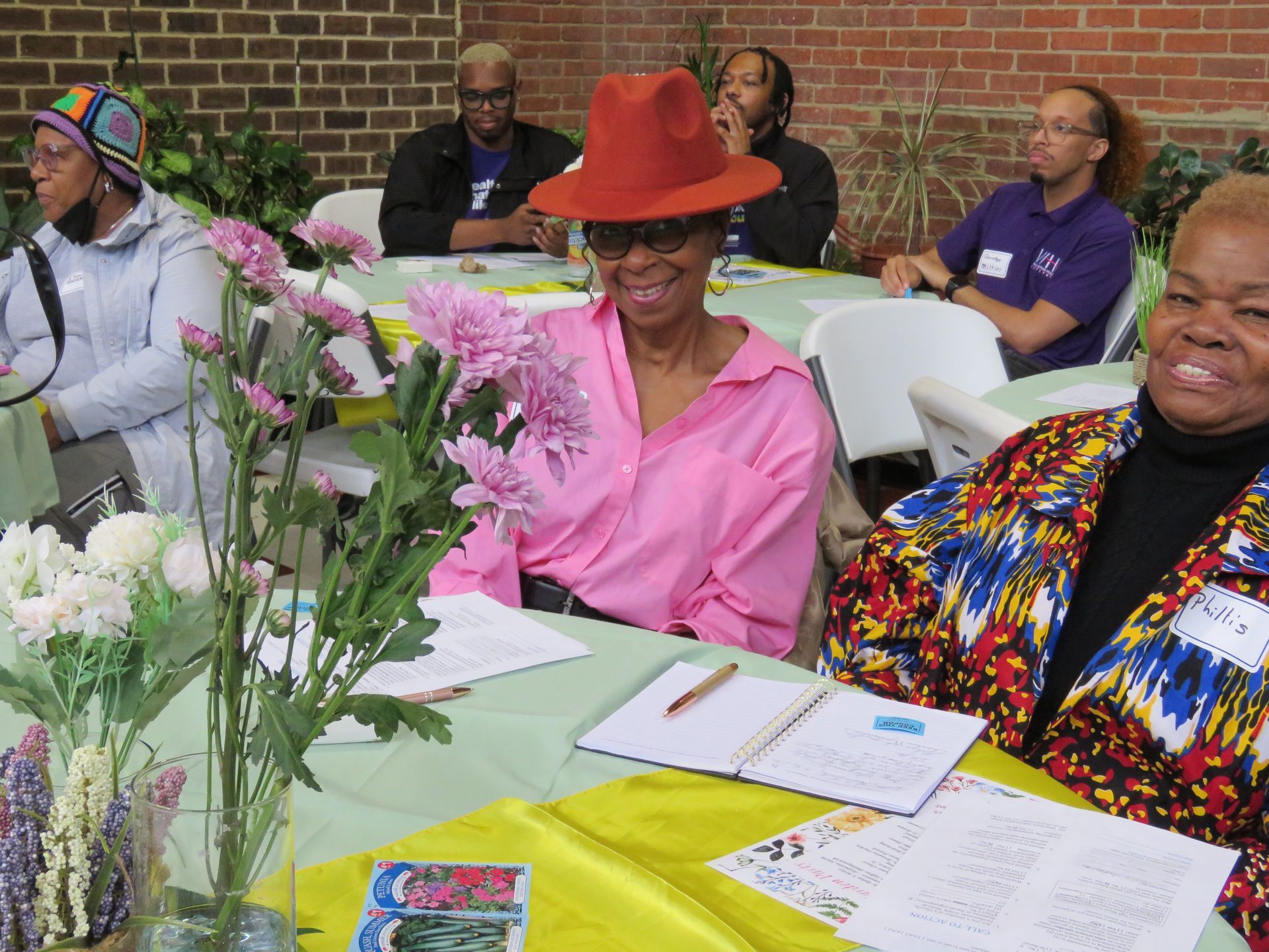 People seated around tables with flowers; one woman wears a pink shirt and hat, smiling.