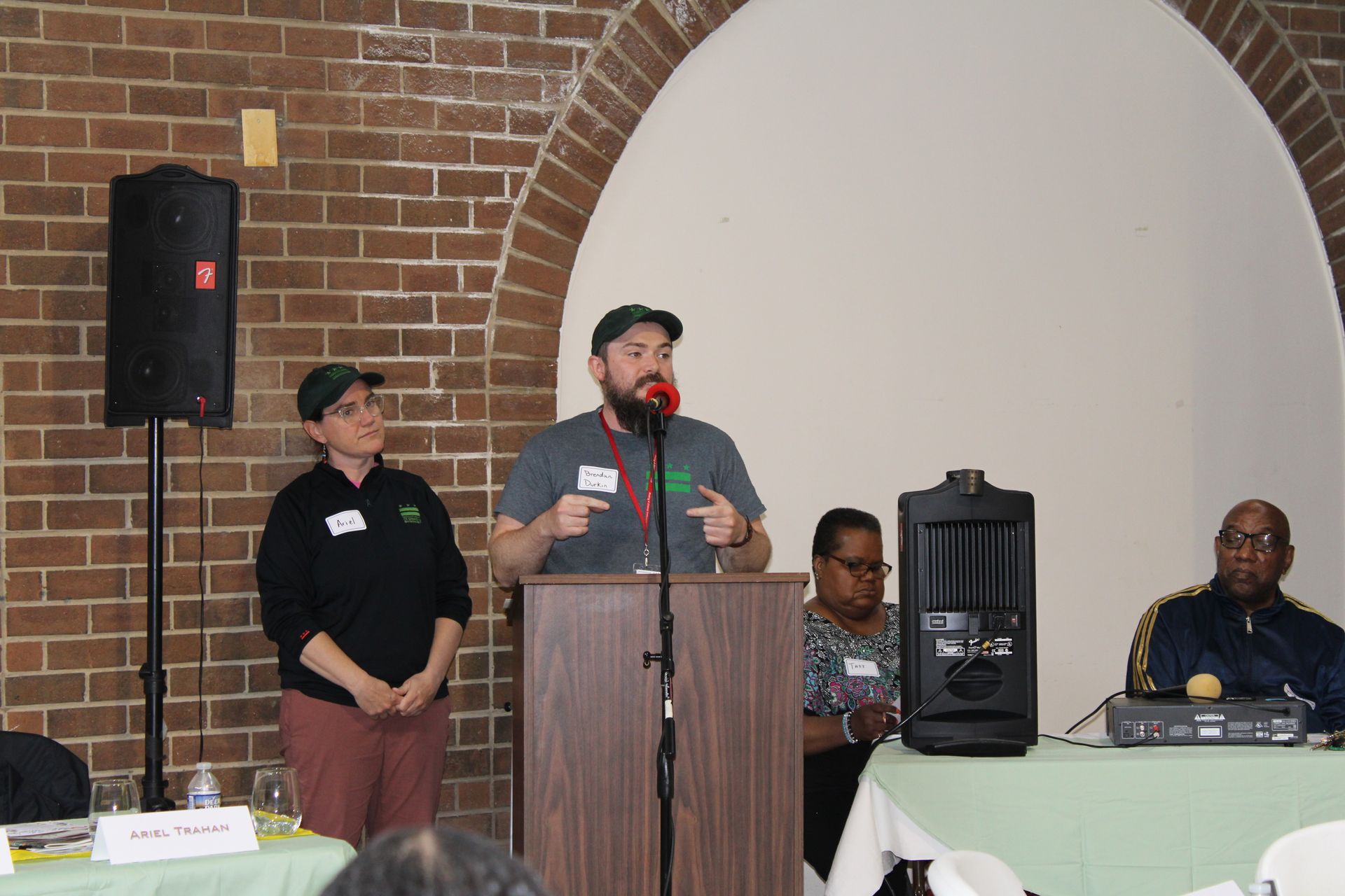 Man speaking at a podium with two others at a meeting; brick wall backdrop.