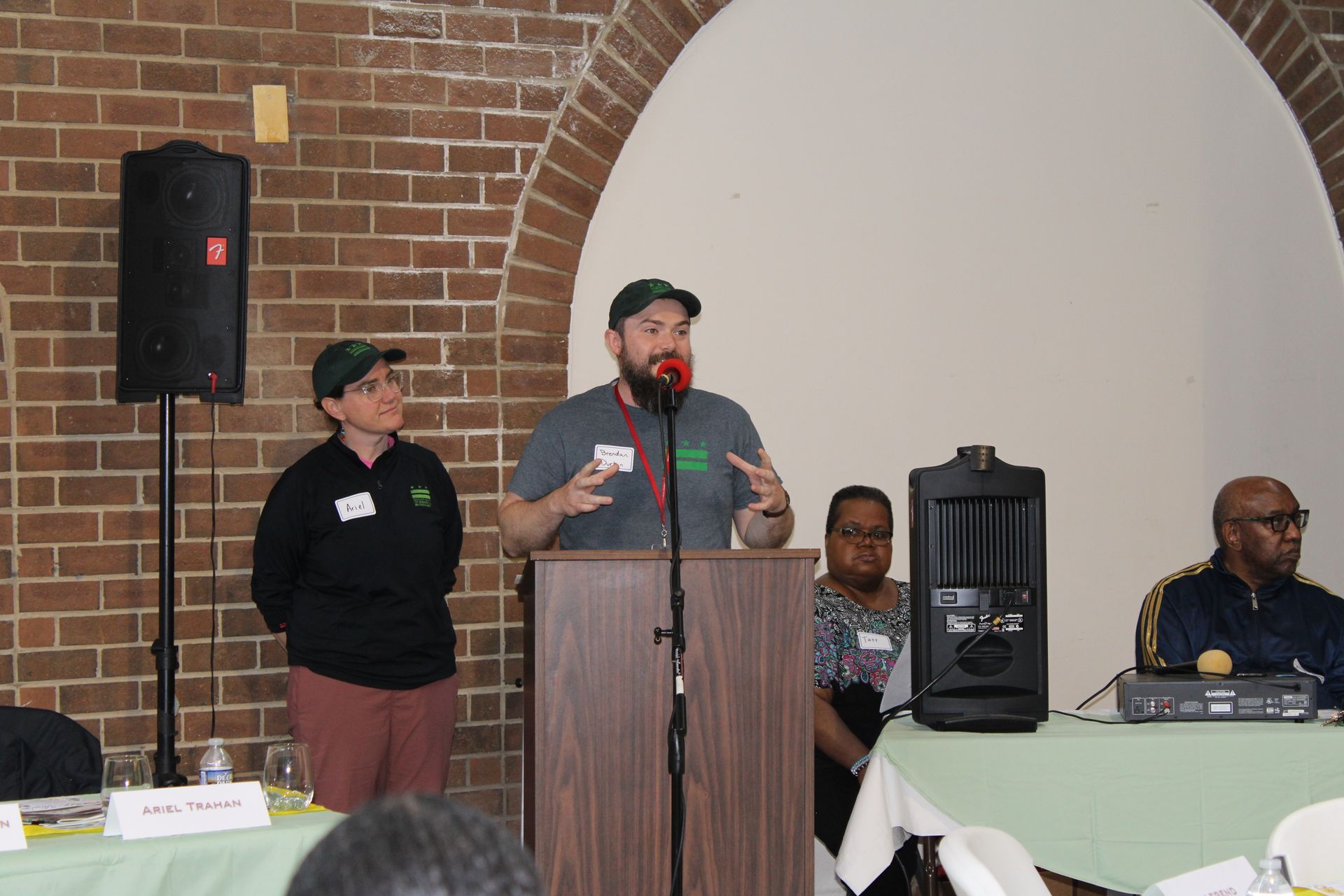 Man speaking at a podium with two people beside him, panel discussion. Brick wall background.
