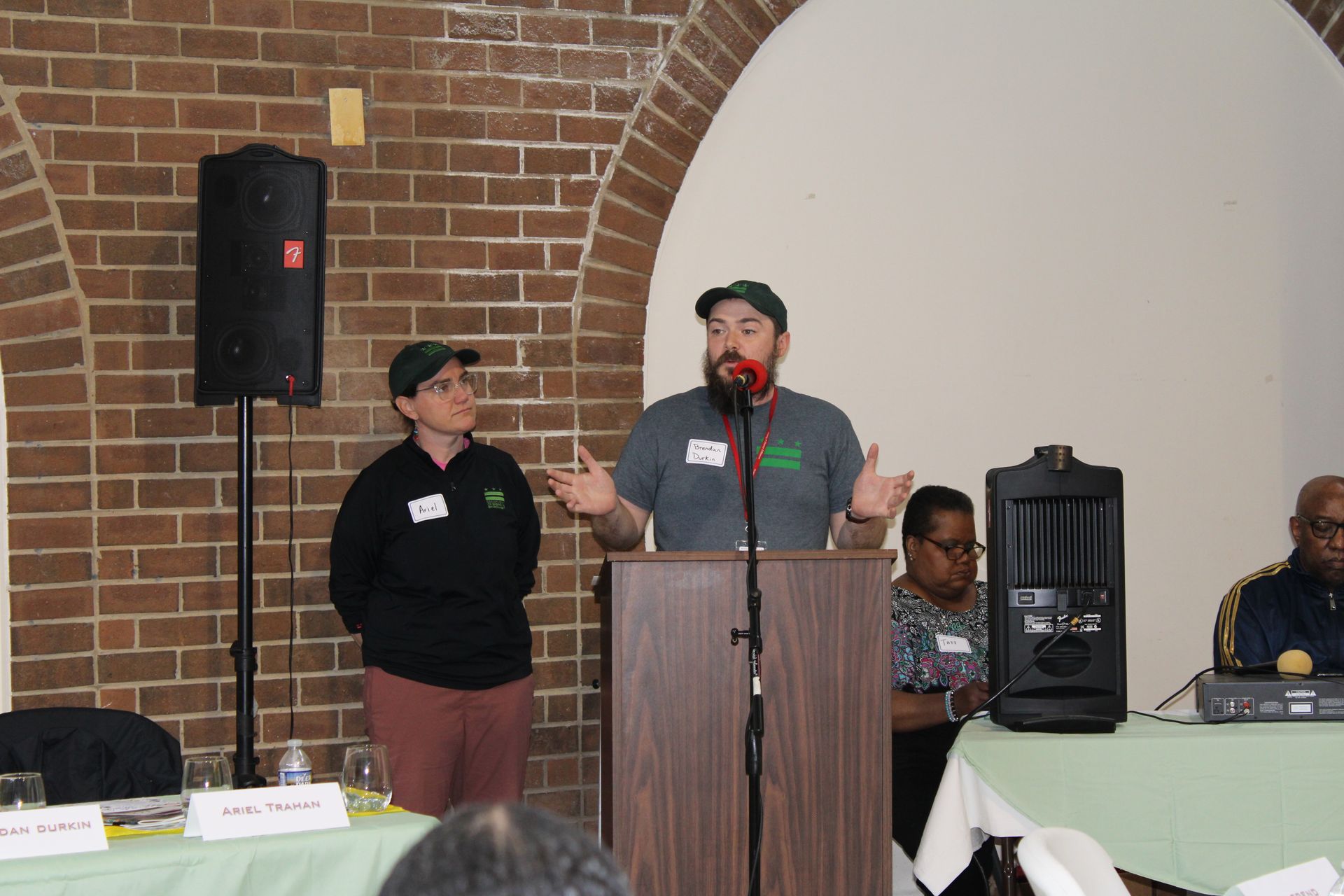 Two people speaking at a podium in front of a brick wall, with additional people seated at a table.