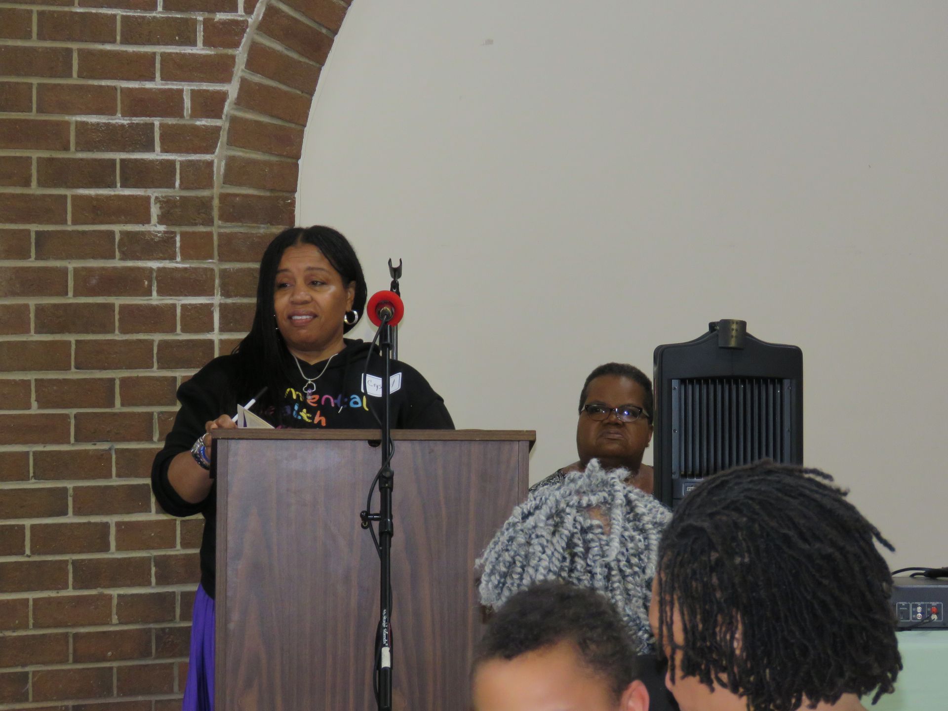 Woman speaking at podium; brick wall backdrop; two people seated nearby.