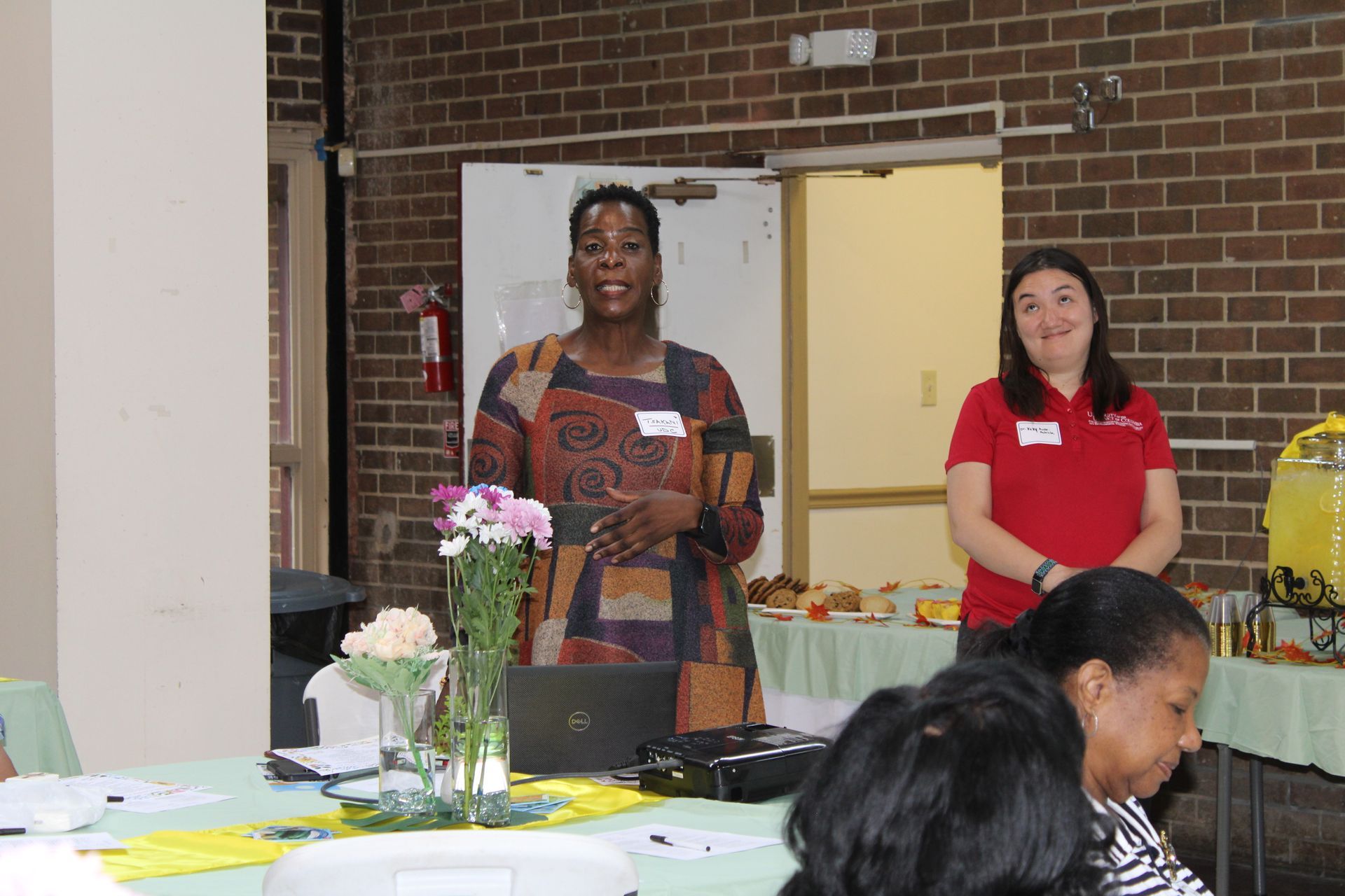Two women at a table. One speaks while the other smiles. Table has flowers, food, and laptop. Brick wall backdrop.