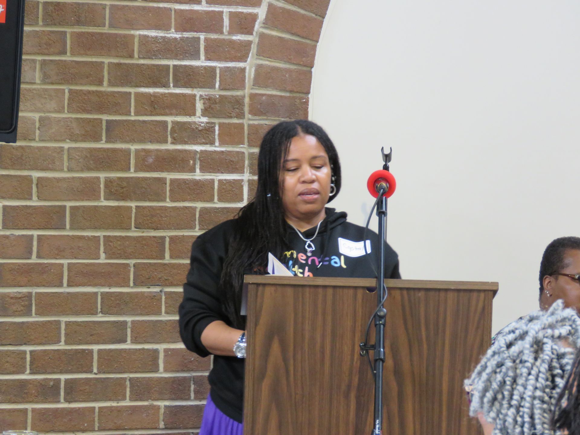 Woman speaking from a podium in a brick-walled room. She wears a black hoodie and purple skirt.
