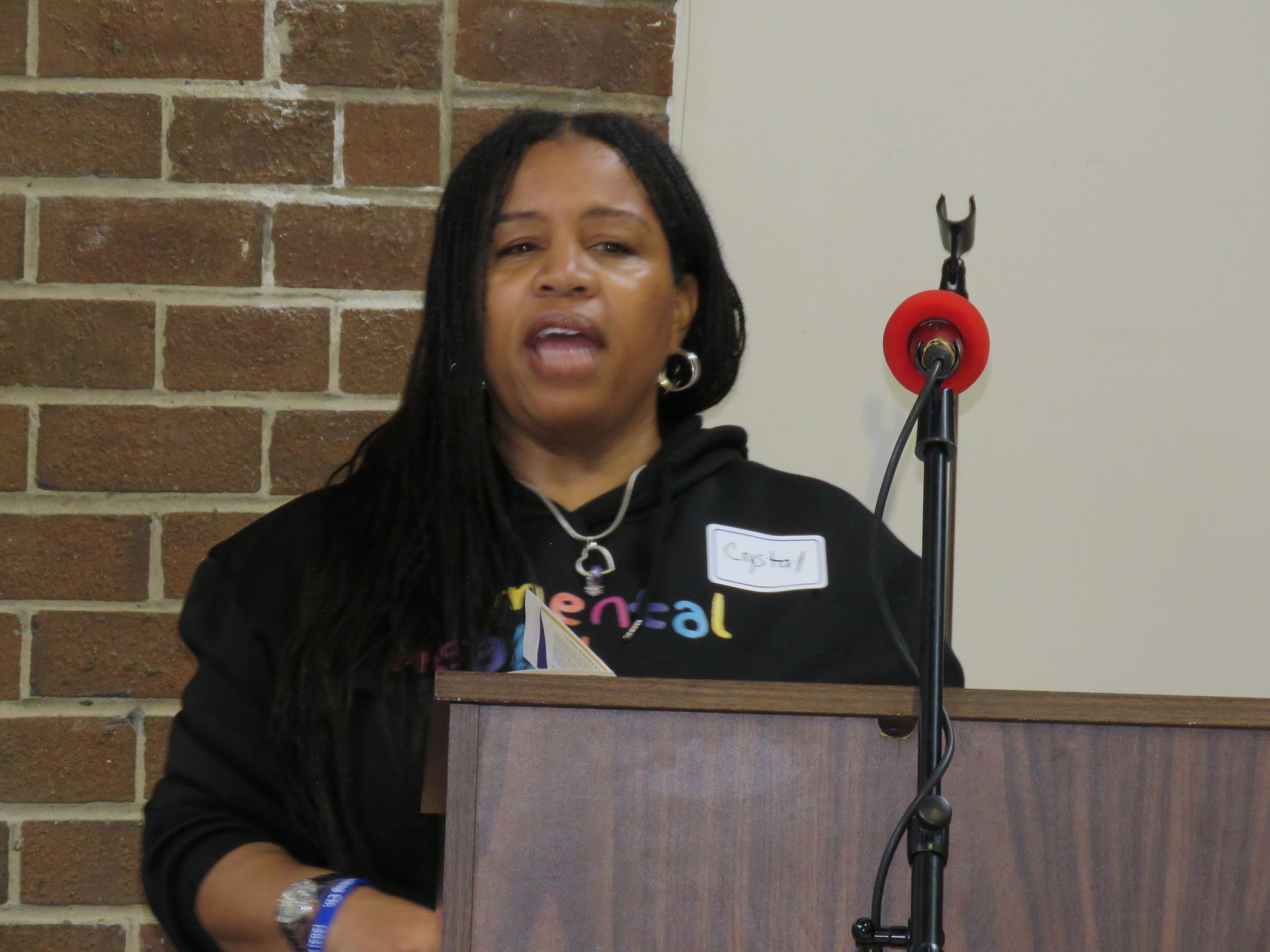 Woman speaking at a podium, wearing a black sweatshirt with a colorful design. Brick wall background.