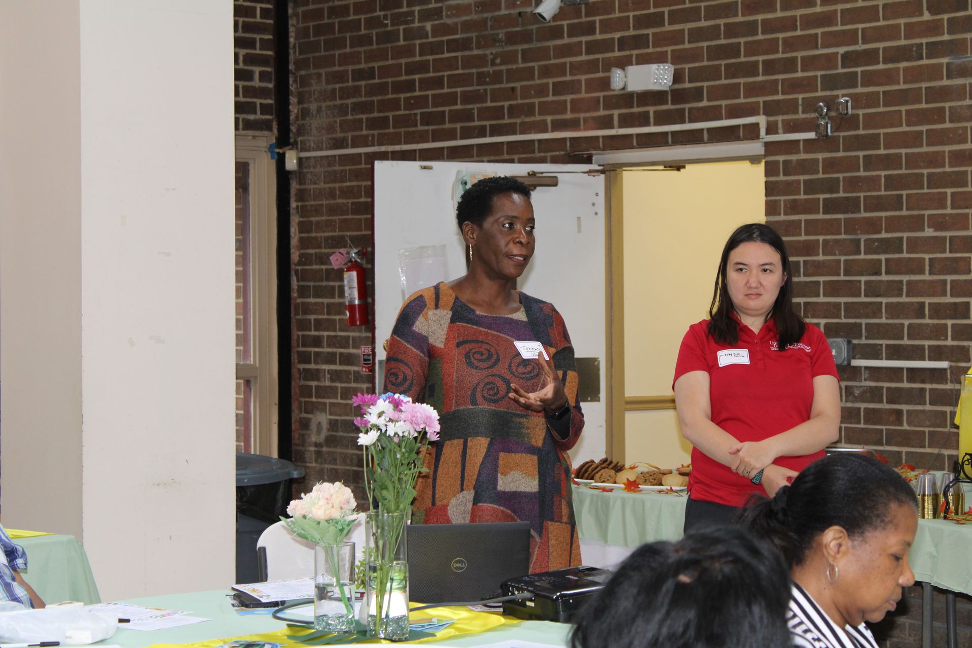 Woman speaking at a table with a laptop, another woman standing beside her, in front of a brick wall.