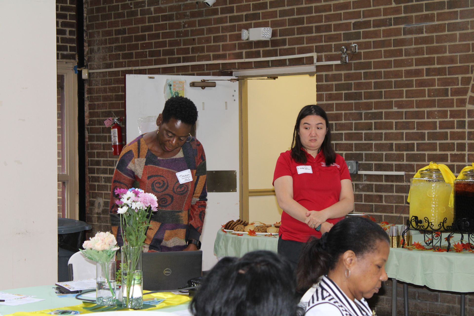 Two women stand at a table during a presentation. One looks down, the other stands.