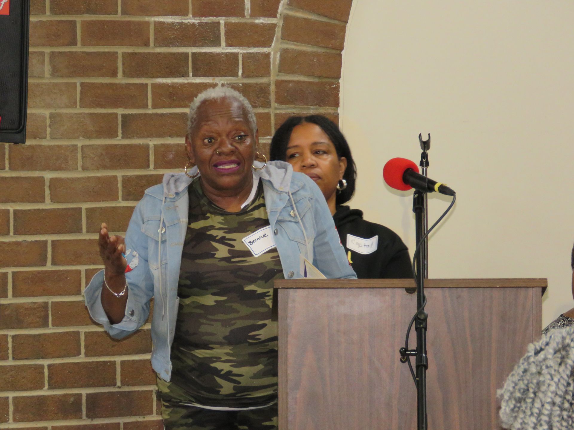 Woman speaking at a podium, gesturing with her hands. Another woman stands behind her. Brick wall in the background.
