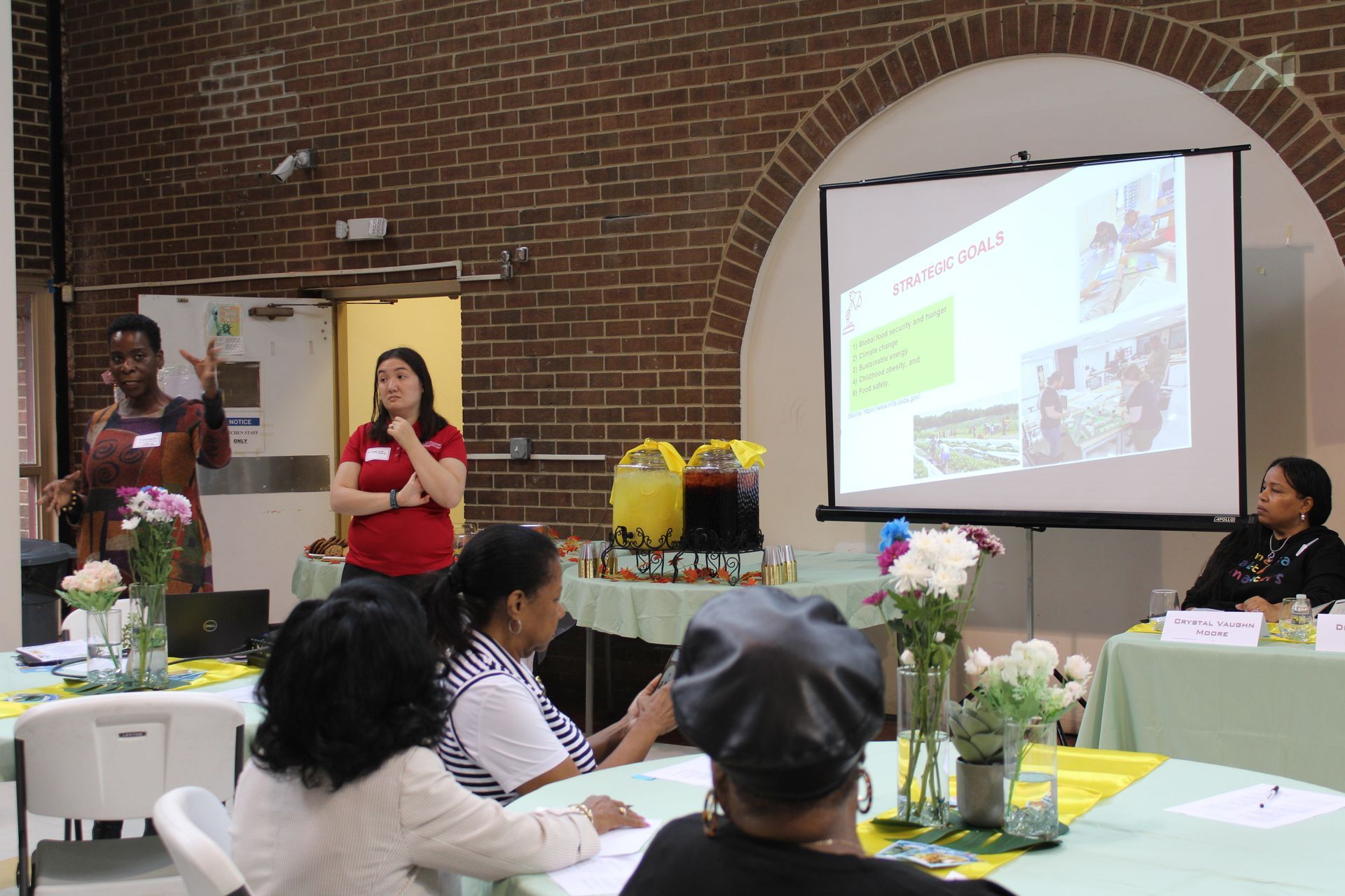 People at a presentation with a projector screen. Two speakers, audience, and refreshments in a room with brick walls.