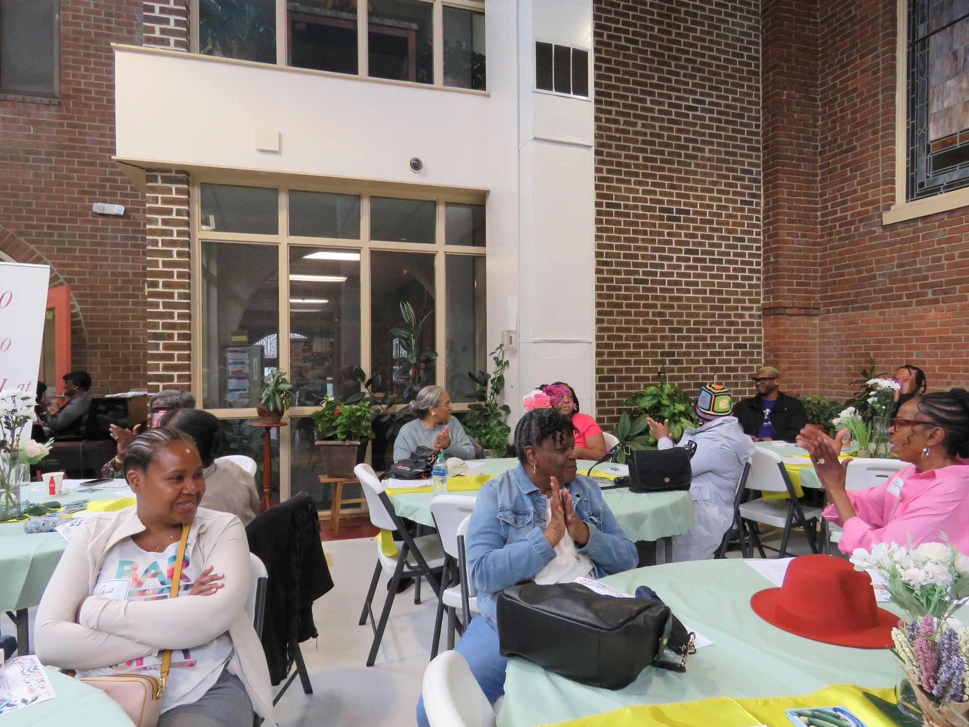 People seated at tables in a room with brick walls. Some are clapping, others listening. Sunlight streams through a window.