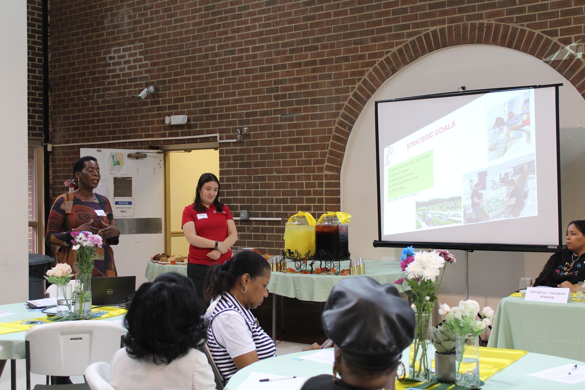 Two women present to an audience; slideshow on a screen. Tables are set with flowers.