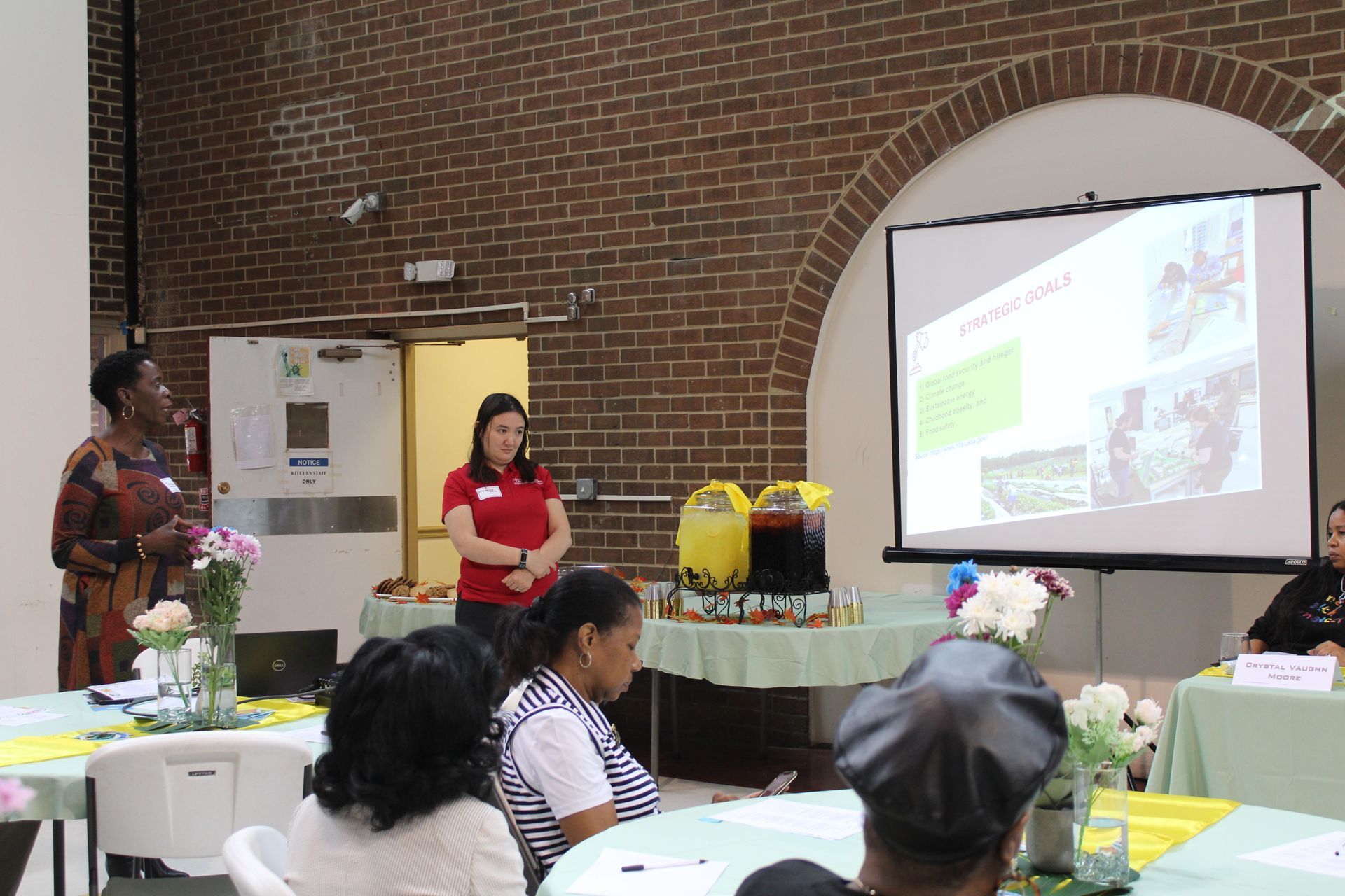 Two women present to an audience, projected slides shown, event in a room with brick wall.