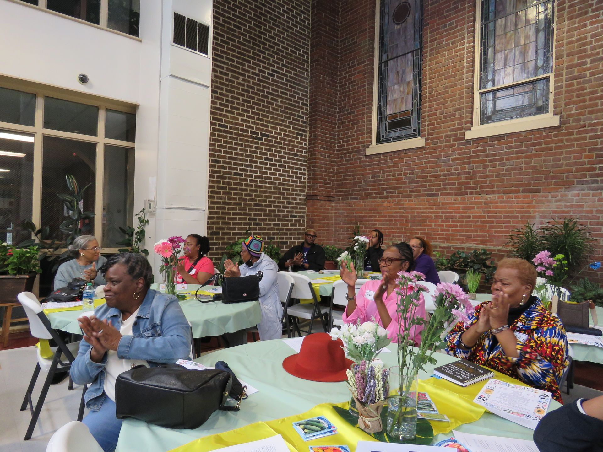 People seated at tables with flowers, clapping. Brick wall and arched windows in background.