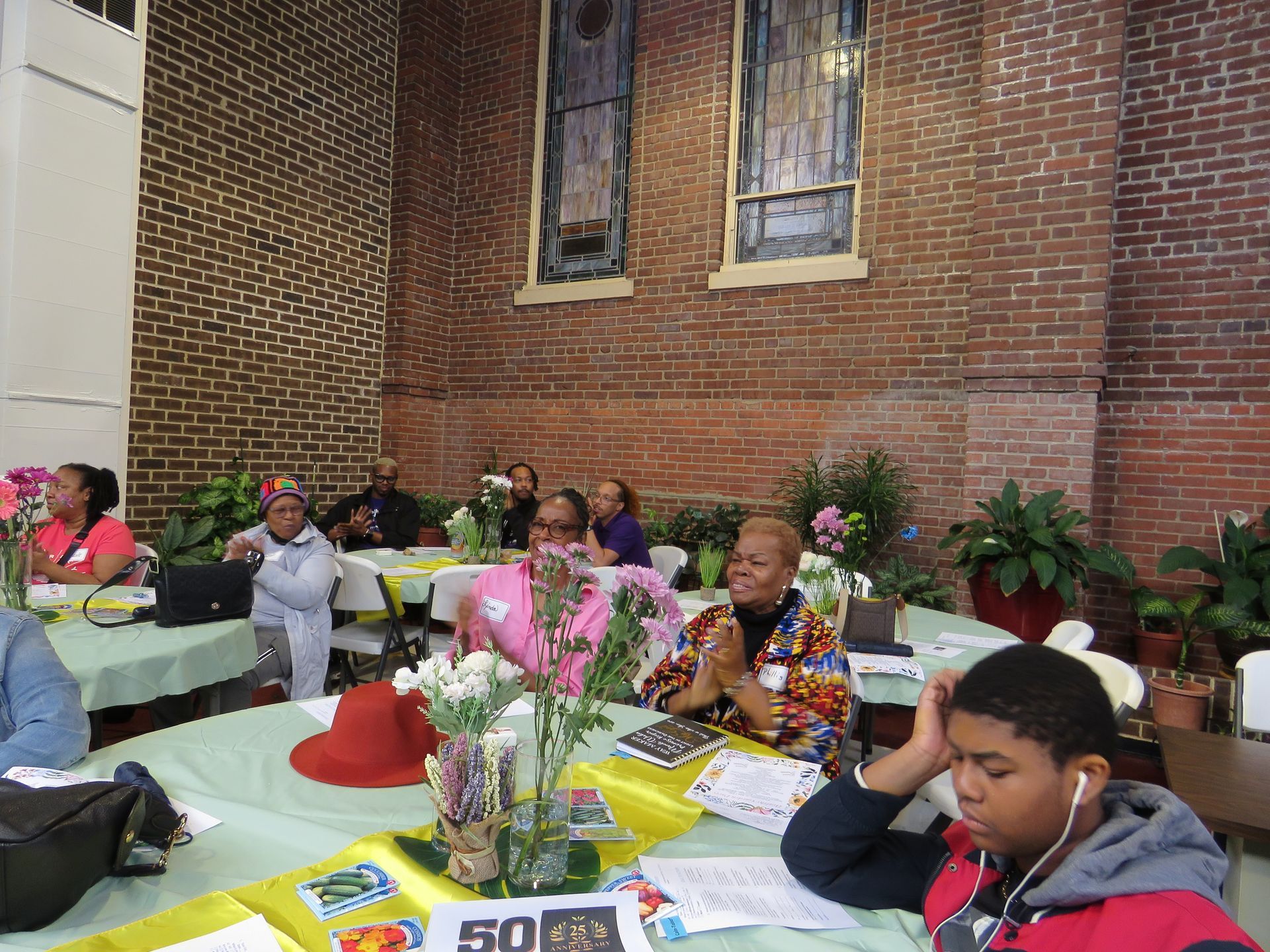 People seated at tables in a room with brick walls, possibly an event. Some are wearing hats and there are flowers.