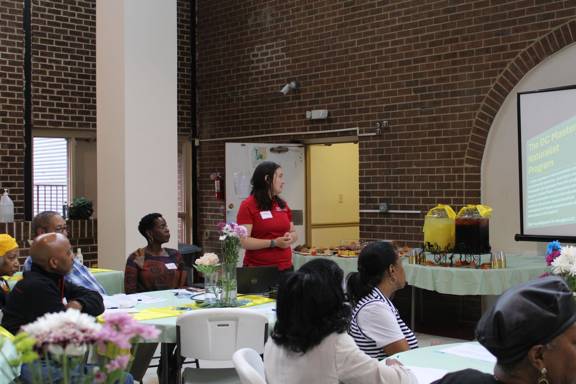 Woman in red shirt presenting to group at table; projector screen, brick wall background.